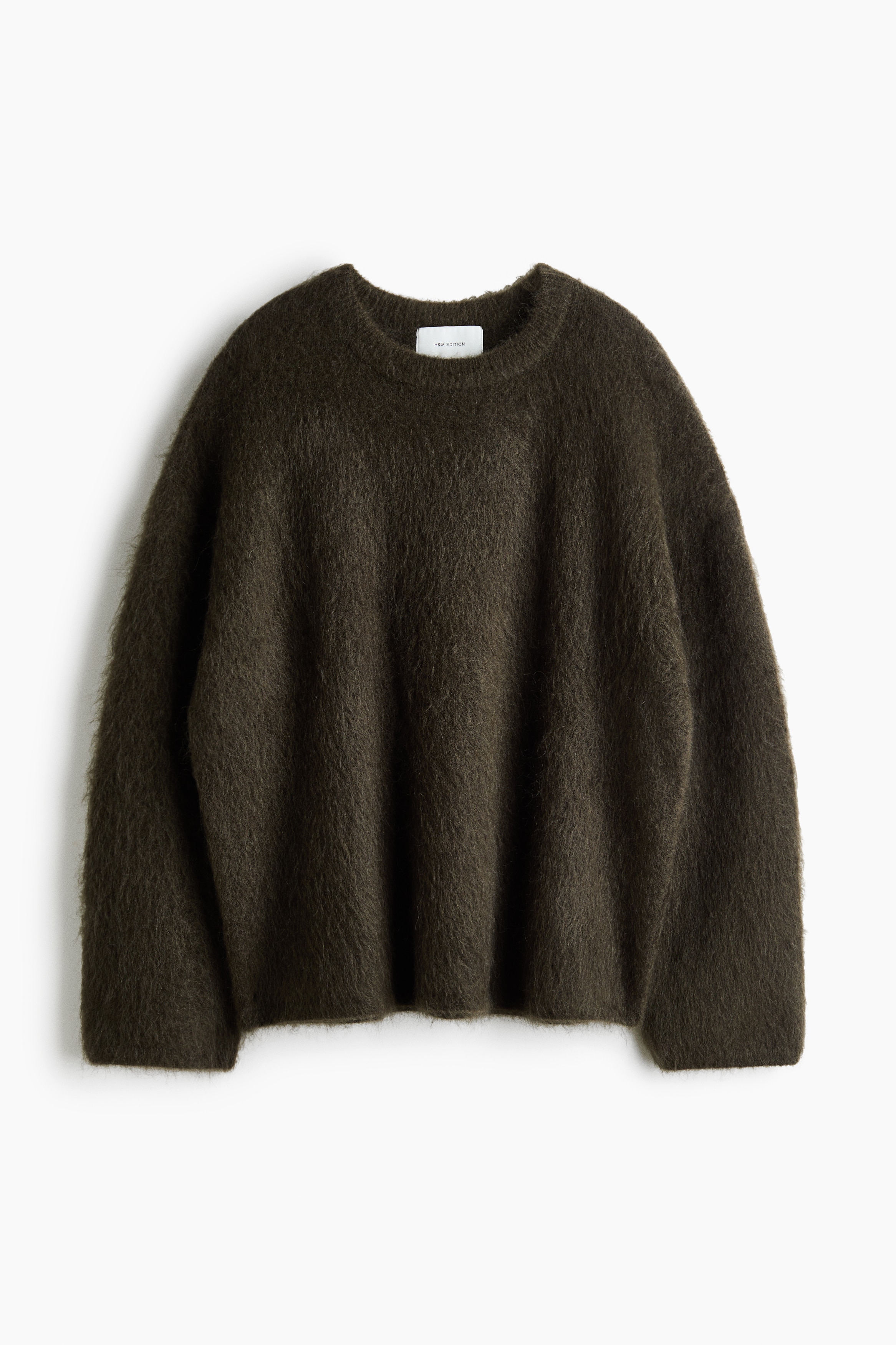 An oversized, dark brown fuzzy knit sweater is being worn by the model, who is holding the fabric of one long sleeve. The sweater features dropped shoulders, a ribbed crew neckline, and is layered over a white top visible at the collar and cuff.