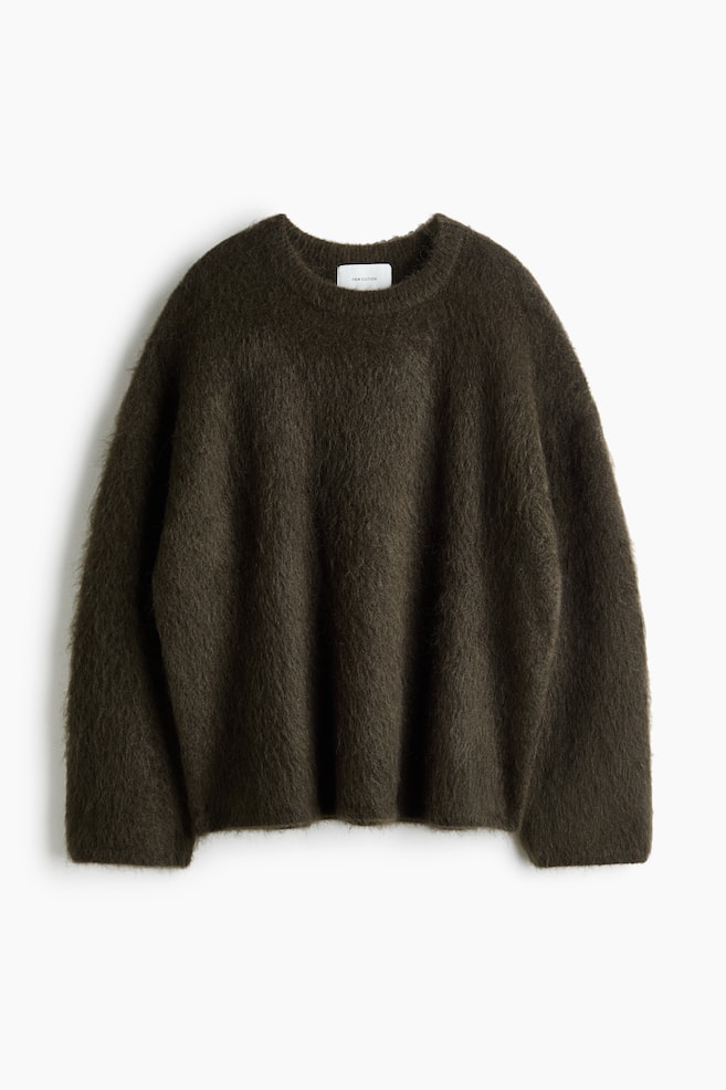 Ein oversized geschnittener Pullover aus mohairhaltigem Strick in dunklem Olivgrün hängt mit der Vorderseite nach vorne und zeigt seine flauschige, gebürstete Textur sowie seine entspannte Silhouette. Er verfügt über überschnittene Schultern, einen gerippten Rundhalsausschnitt und lange, lockere Ãrmel. - 1