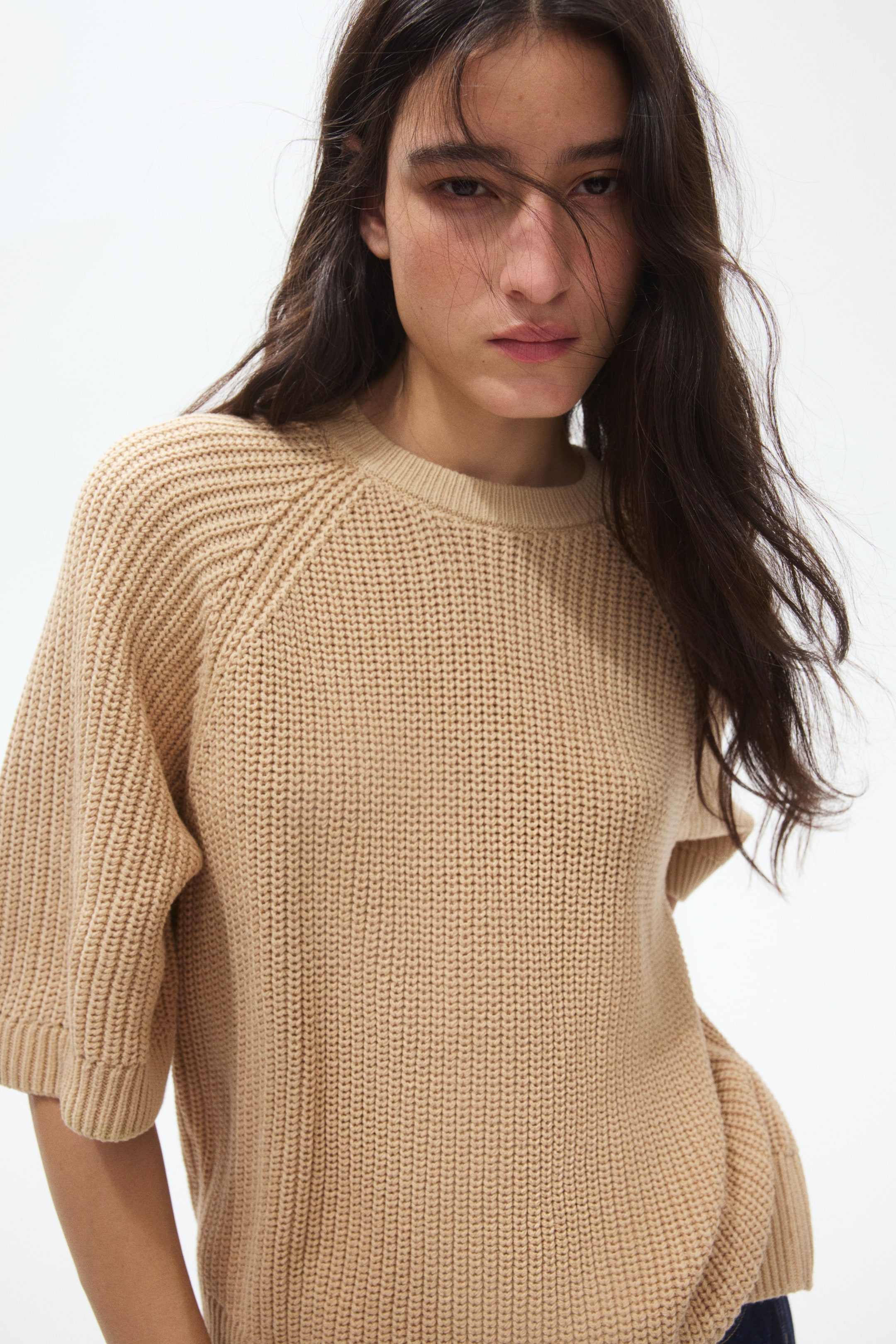 View larger image: A beige knit jumper features a crew neck and short raglan sleeves with ribbed cuffs and ribbed raglan lines. The jumper, crafted with a large gauge stockinette stitch, showcases a relaxed fit from a slight front angle.