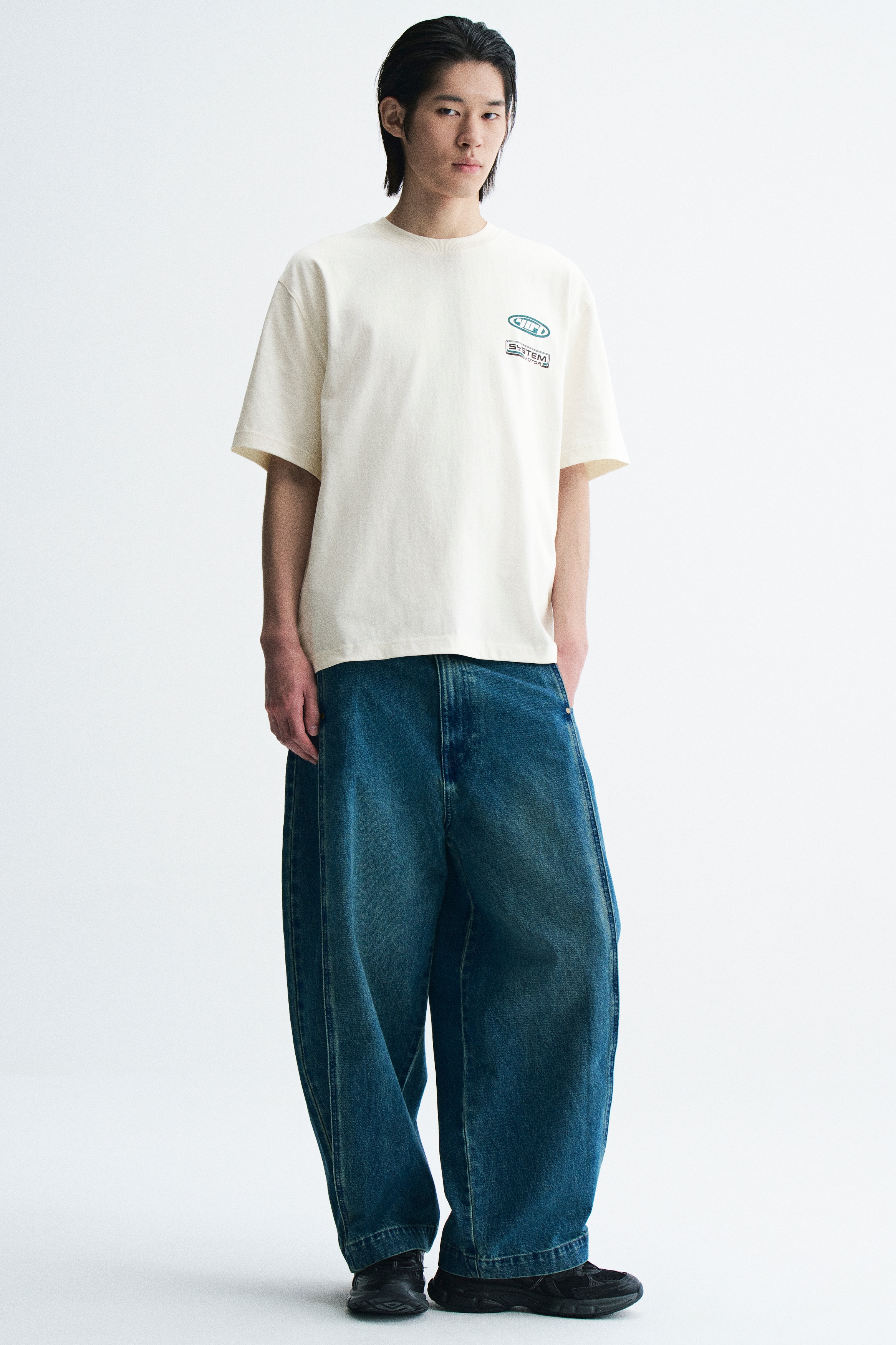 An off-white T-shirt is shown from the front, featuring a ribbed crew neck, short sleeves, and two small graphic designs on the left chest, including a teal oval emblem and a rectangular logo with the text "SYSTEM MOTOR".