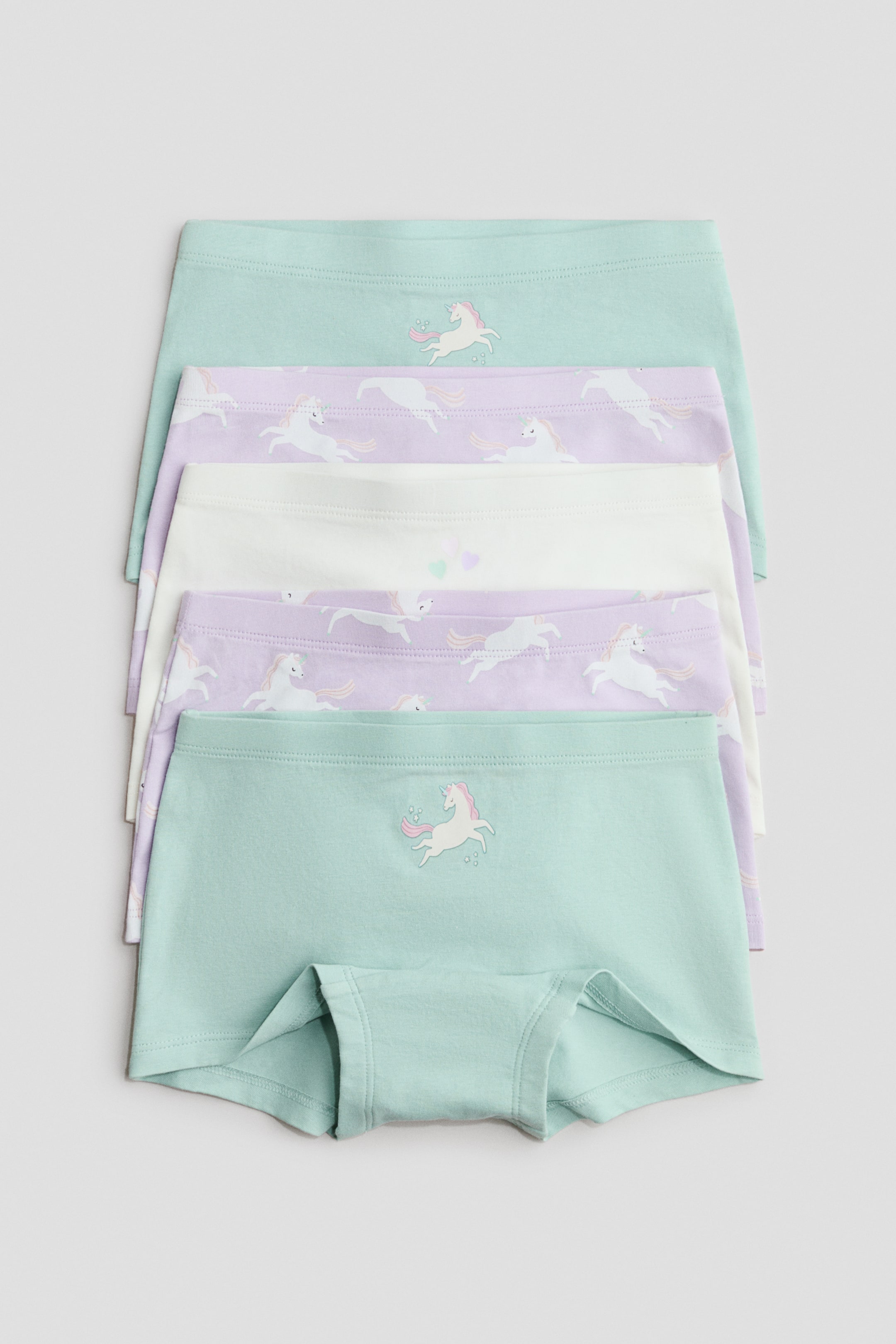 Ver imagen más grande: Cinco calzoncillos de jersey de algodón suave están apilados, con una cintura elástica y varios diseños. Dos calzoncillos son de color verde menta, con un gráfico de unicornio en la parte delantera; dos son de color púrpura claro con un patrón de unicornios por todas partes; y uno es blanco con pequeños corazones, mientras que el calzoncillo inferior también revela un refuerzo forrado en su parte delantera.