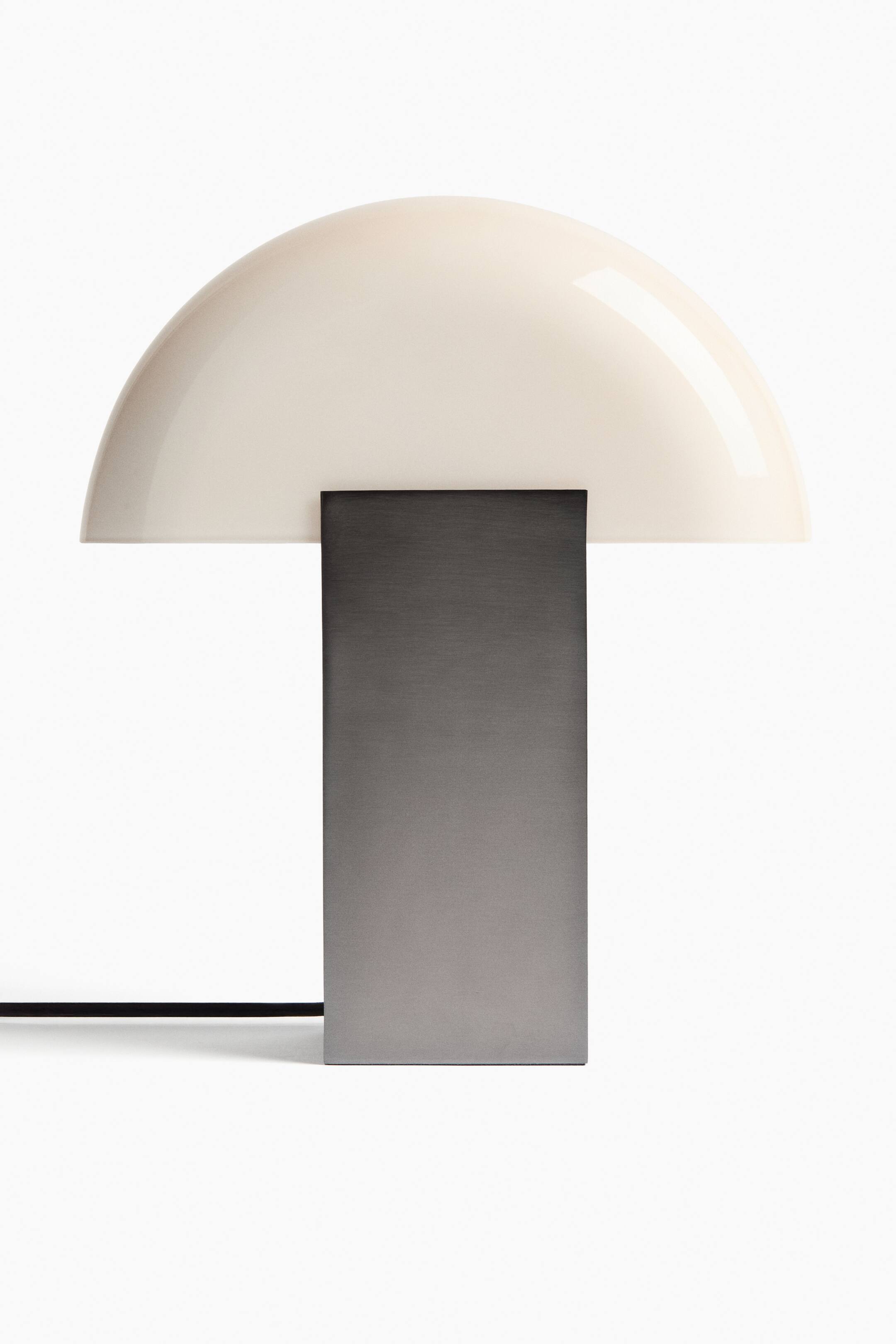 Größeres Bild ansehen: Eine Tischlampe zeigt einen cremefarbenen, gewölbten Schirm, der auf einem flachen, rechteckigen, dunkelgrauen Metallfuß ruht, mit einem schwarzen Netzkabel, das von der linken Seite ausgeht.