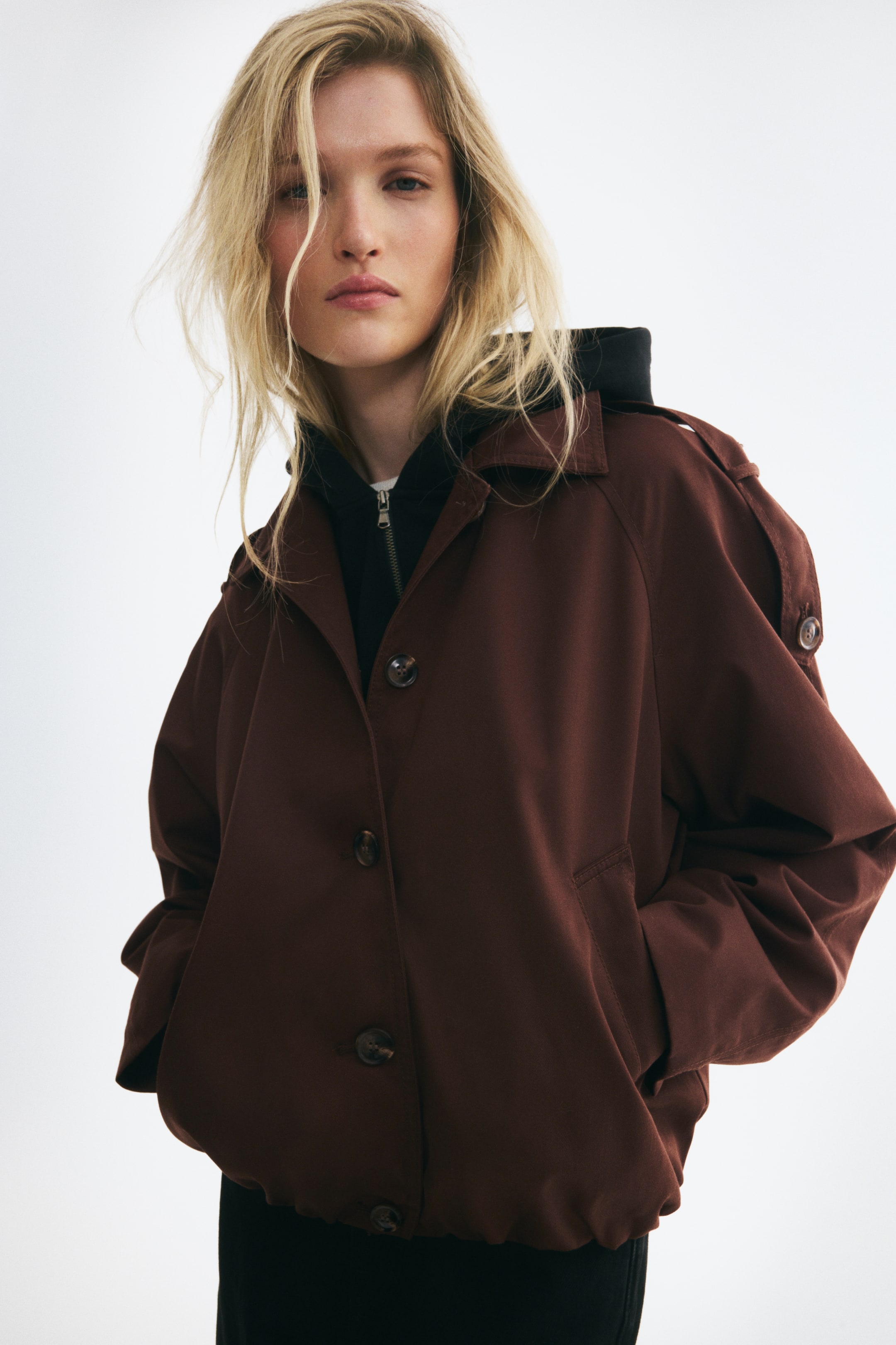 Agrandir l'image: Un blouson oversize en sergé brun foncé est présenté de face, avec une fermeture boutonnée, des manches raglan ornées de pattes d’épaule boutonnées et des poches passepoilées sur les côtés. La veste est portée sur un sweat à capuche noir, mettant en valeur sa silhouette bouffante et son bas élastiqué à effet ballon.
