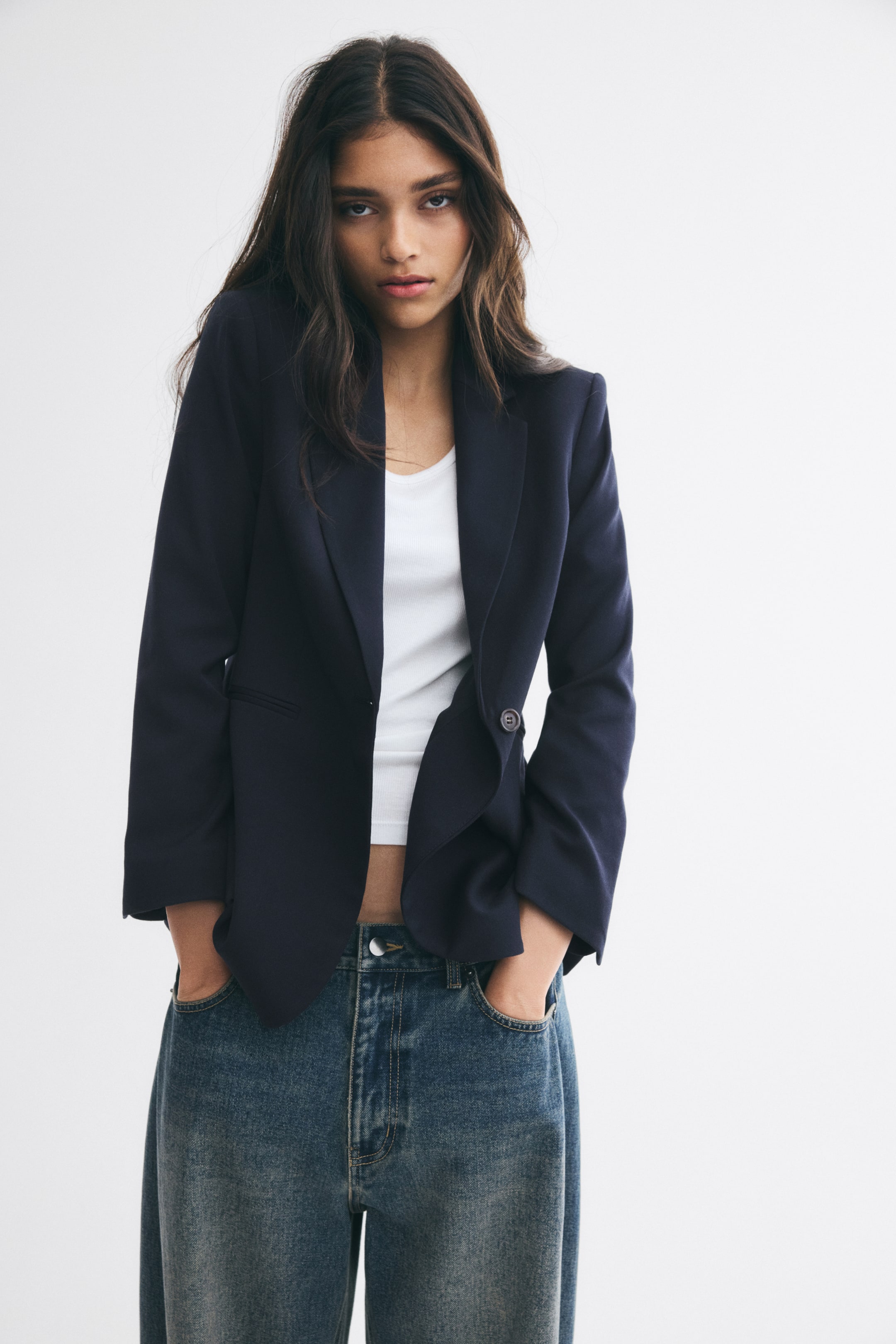 View larger image: A dark blue, single-breasted blazer is worn open by the model over a white tank top and blue jeans. The slim-fit blazer features notch lapels and a single front button, with the model posing with hands in the jeans pockets.