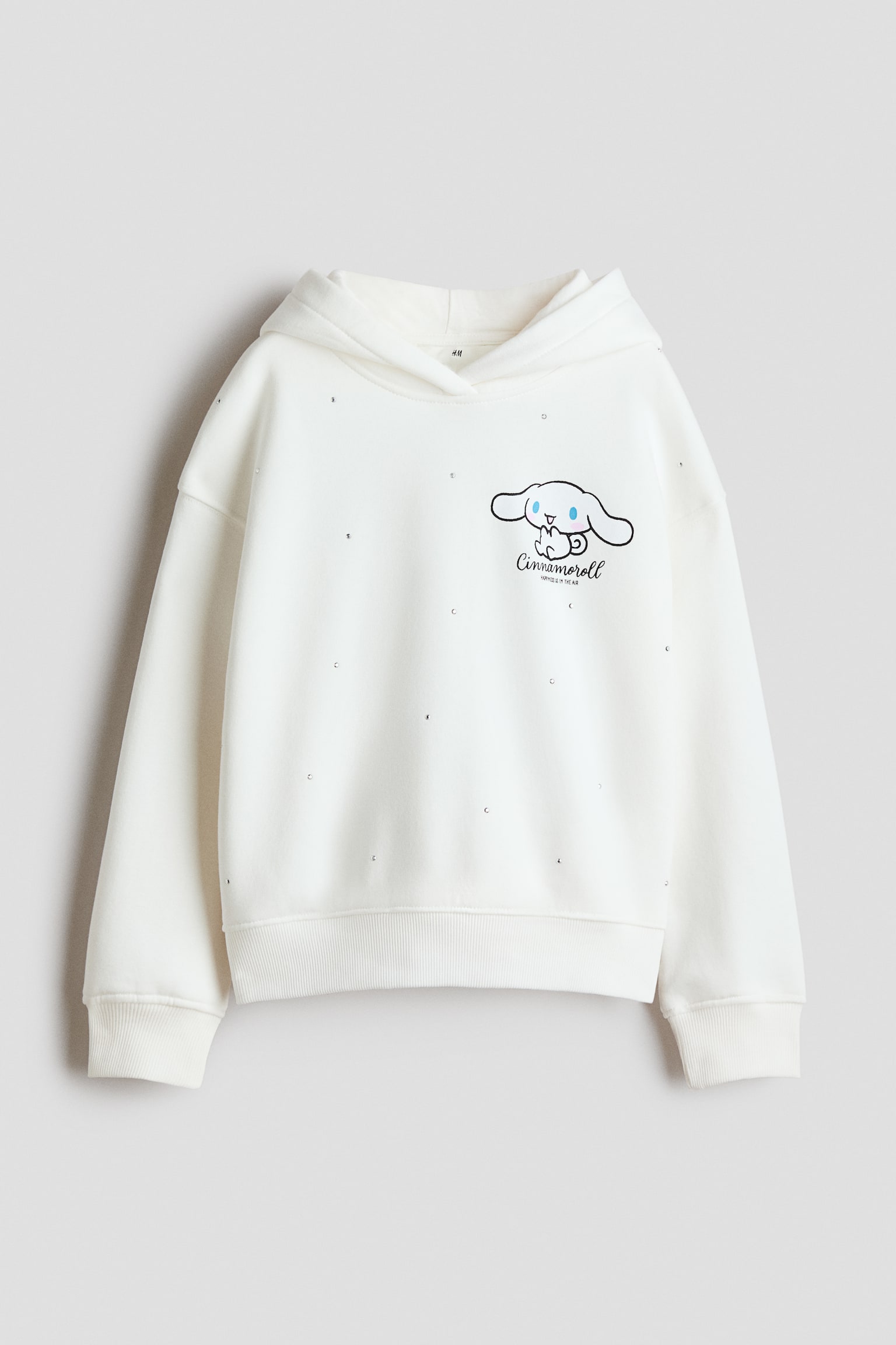 Zepředu je zobrazen oversize mikina s kapucí v odstínu off-white, s potiskem motivu Cinnamoroll na levé straně hrudi a drobnými kamínkovými ozdobami rozmístěnými po celé látce. Design zahrnuje obtočenou kapuci, spuštěná ramena a dlouhé rukávy s žebrovanými manžetami a žebrovaným lemem.