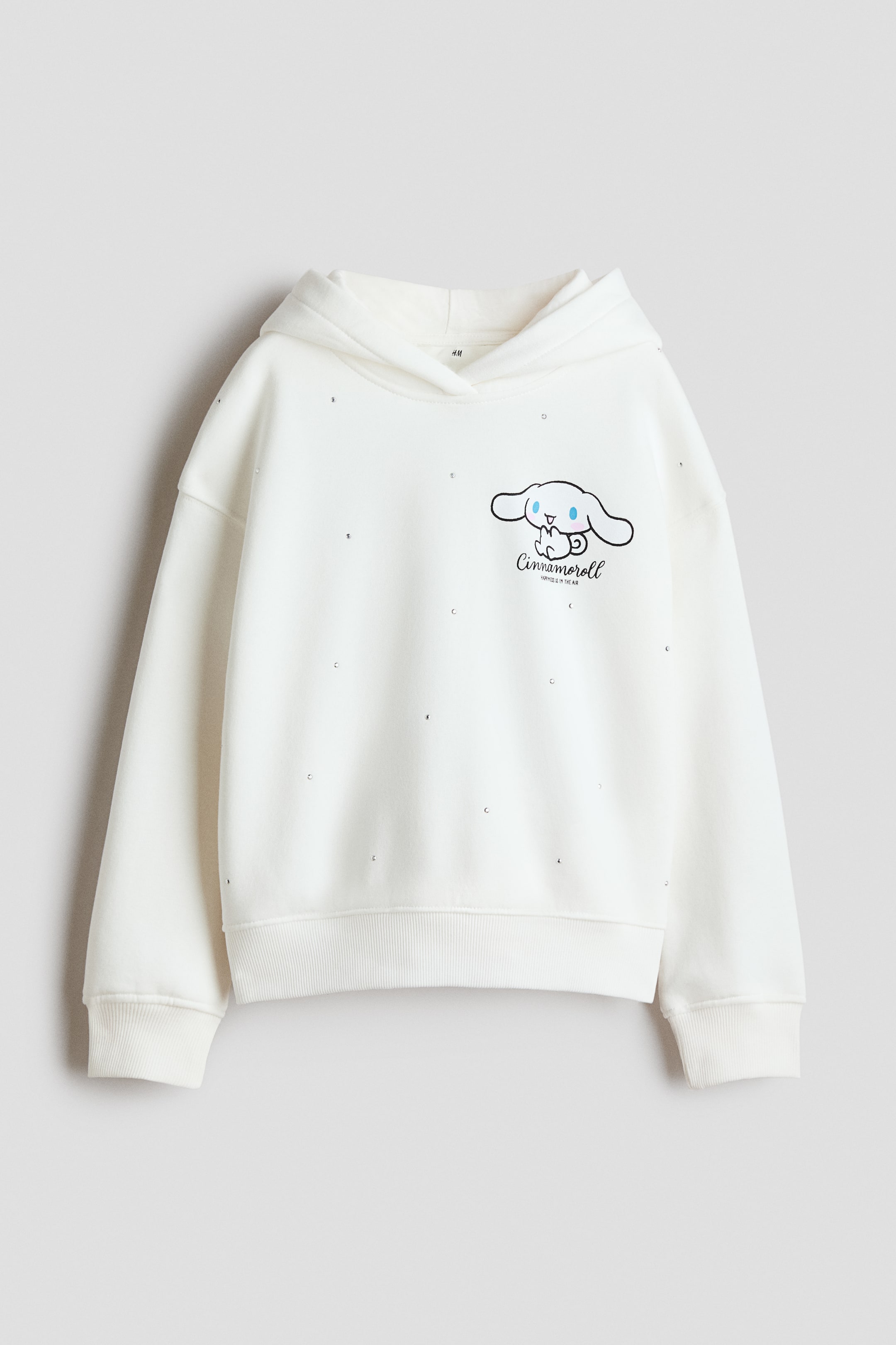 Größeres Bild anzeigen: Ein cremefarbenes Oversize-Hoodie wird von vorne gezeigt und verfügt über einen auf der linken Brust aufgedruckten Cinnamoroll-Print sowie kleine Strassverzierungen, die über den Stoff verteilt sind. Das Design umfasst eine Wickelkapuze, überschnittene Schultern und lange Ärmel mit Rippbündchen sowie einen Rippbund am Saum.