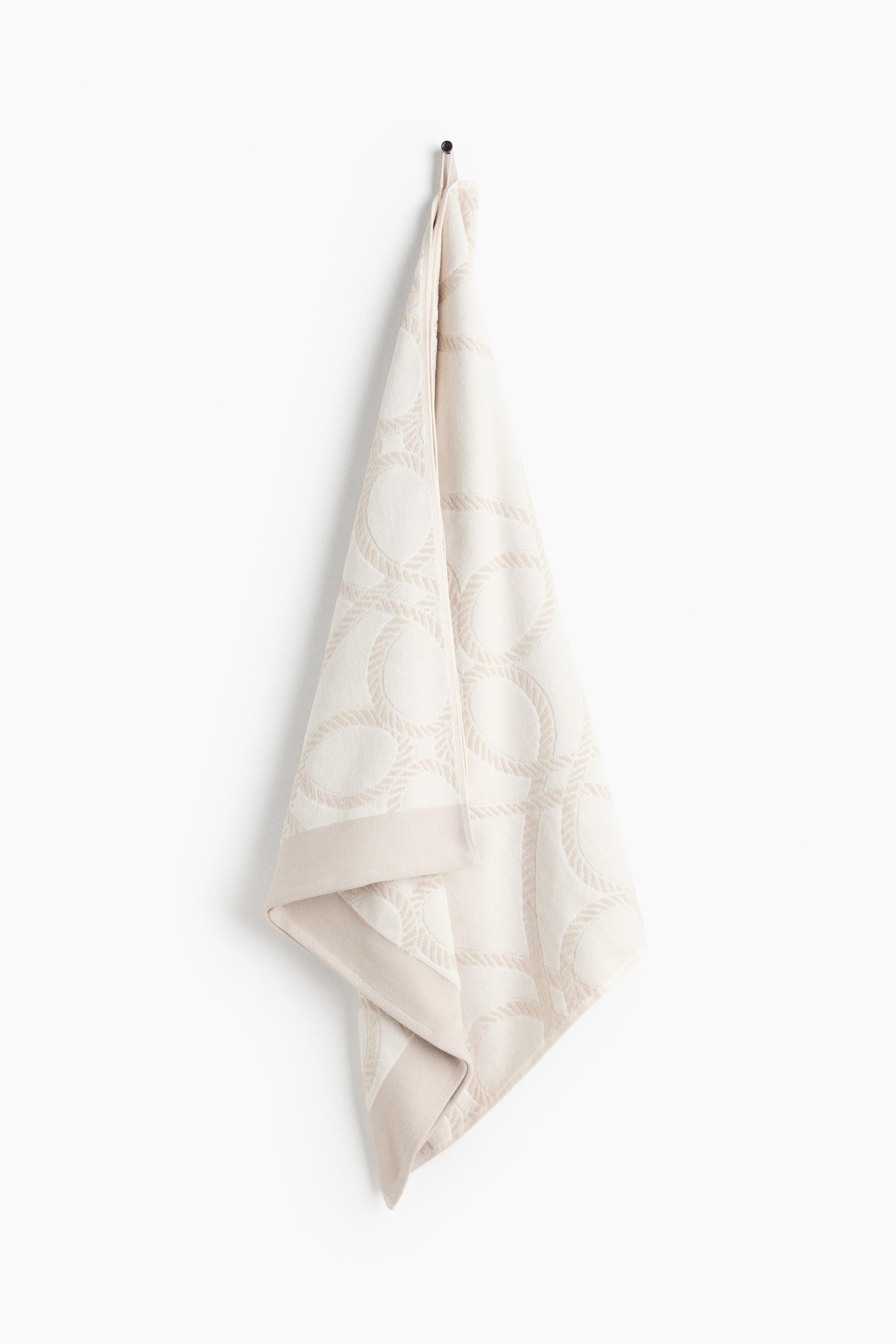 View larger image: The beach towel is suspended, showcasing its velour front crafted from soft cotton. It boasts a cream base adorned with a jacquard pattern of interlocking rope circles, complemented by a solid border in a beige accent hue.