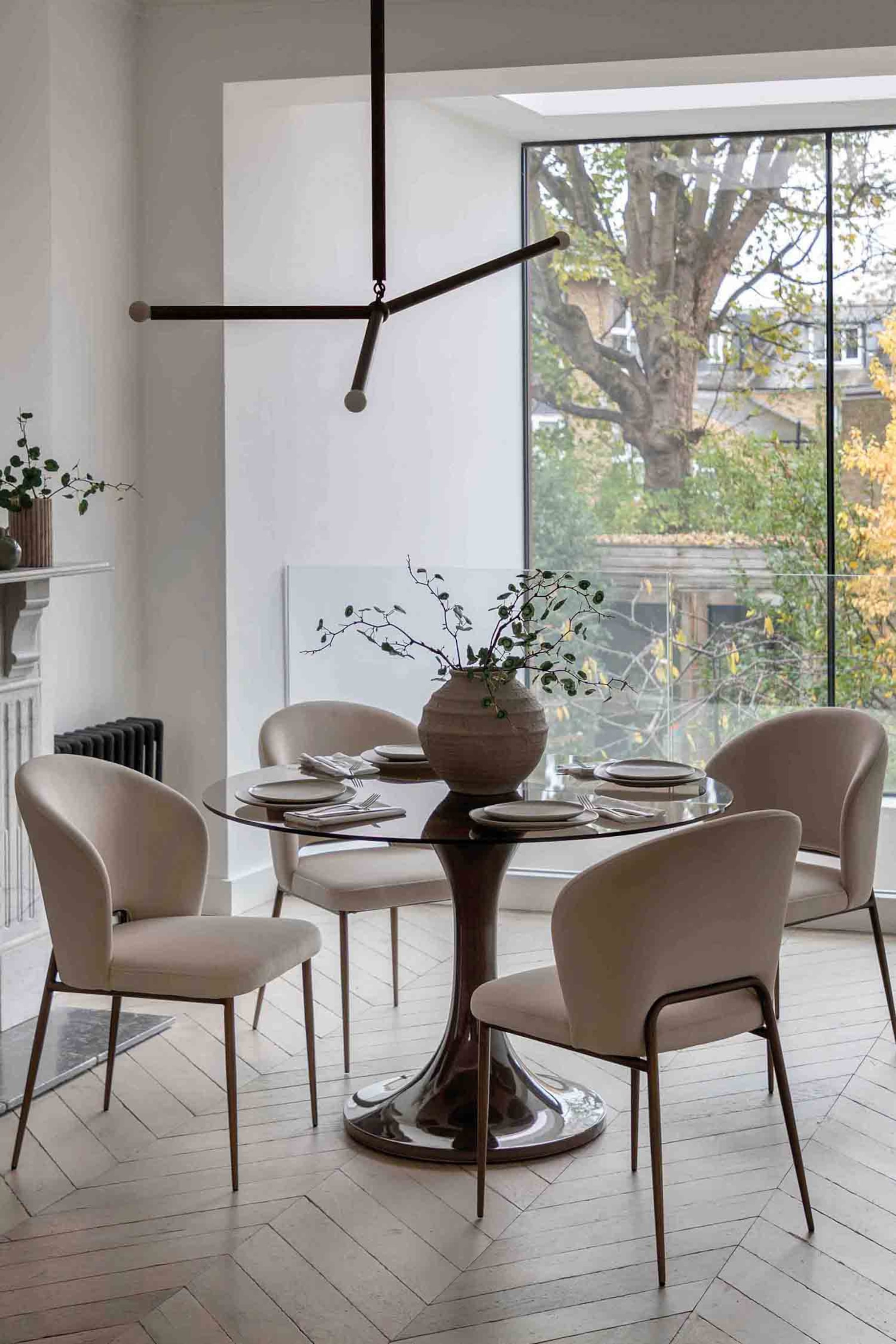 View larger image: A round dining table featuring a dark glass top and a polished metallic, flared pedestal base. Four light beige upholstered dining chairs with curved backs and slender dark metallic legs surround the table, which is set with plates, cutlery, and a centrepiece vase.