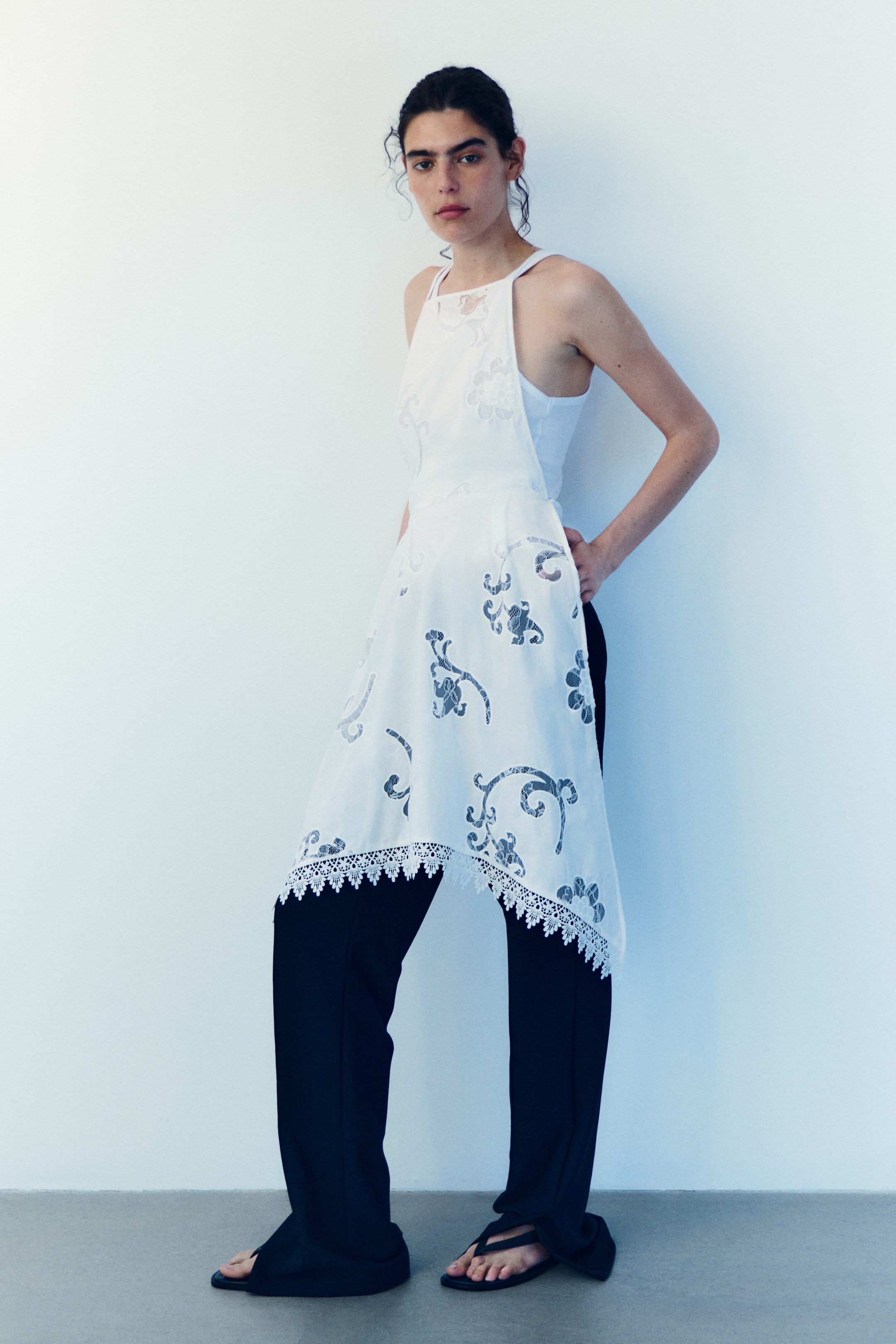 View larger image: A white overlay garment featuring a halter-style neckline, intricate floral cutouts revealing a dark pattern underneath, and an asymmetric hemline finished with white lace trim, worn over a white tank top and black pants.