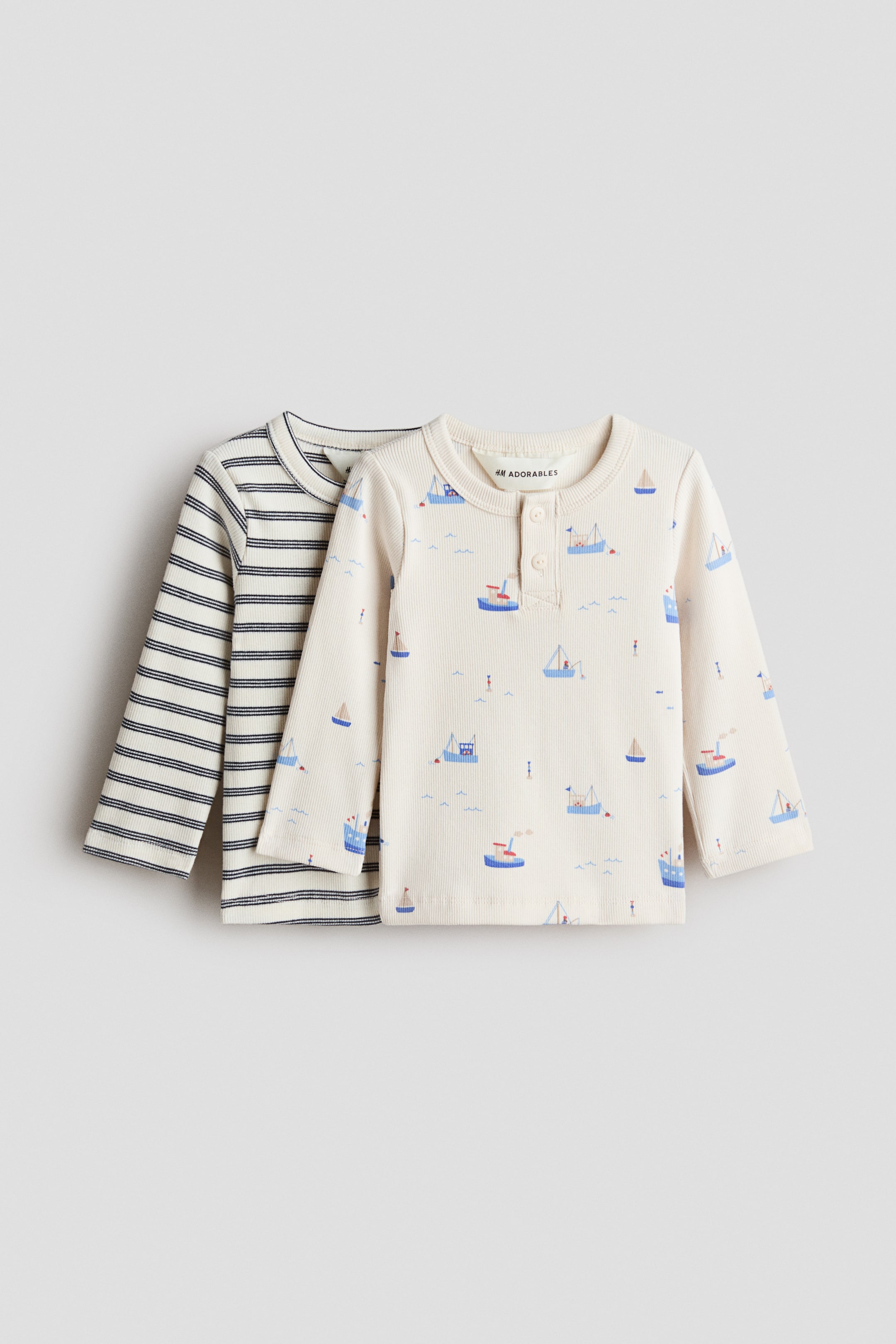 Ver imagen más grande: Dos camisetas de punto acanalado con mangas largas de H&M Adorables están colocadas planas, mostrando sus frentes; la que está al frente es de color blanco hueso con un patrón de velero azul y presenta un cuello redondo con borde acanalado y una solapa de dos botones. Detrás de ella, se ve parcialmente una segunda camiseta de punto acanalado, que muestra una base crema con finas rayas horizontales azul marino y un cuello redondo similar.