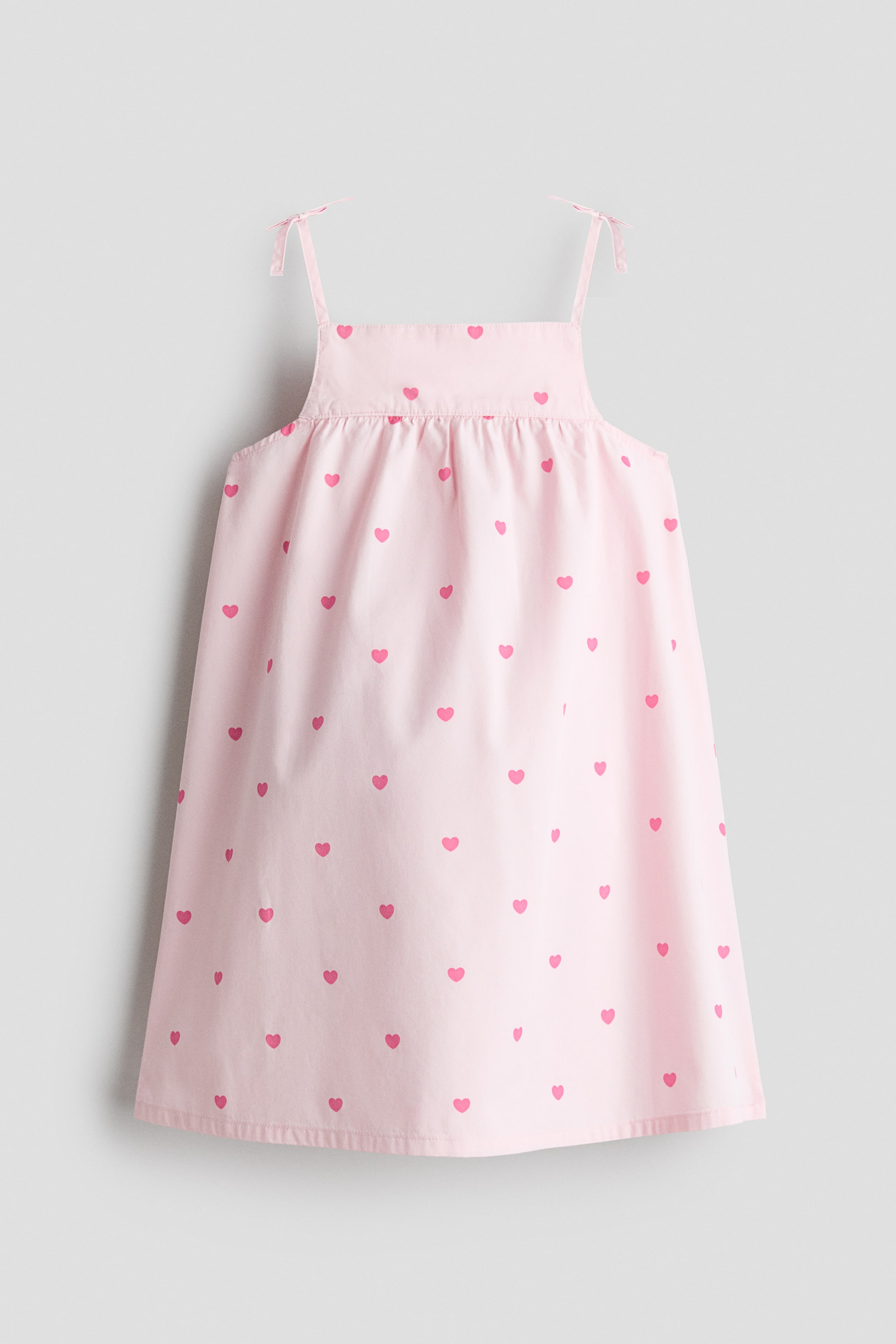 View larger image: A light pink A-line dress with an all-over pattern of small pink hearts is shown from the front. The dress features a straight neckline, narrow shoulder straps with decorative bows, and a gathered seam below the front yoke that adds volume to the flared skirt.