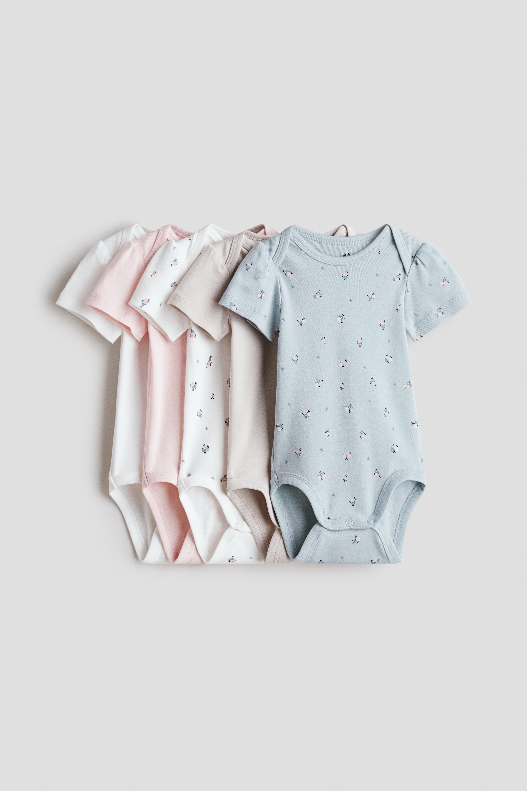 View larger image: A set of five short-sleeve bodysuits is displayed, all featuring lapped shoulders and snap closures at the crotch. The bodysuits are available in solid white, pink, and beige, and in white and light blue with a small floral pattern, with the light blue bodysuit facing forward in the foreground.