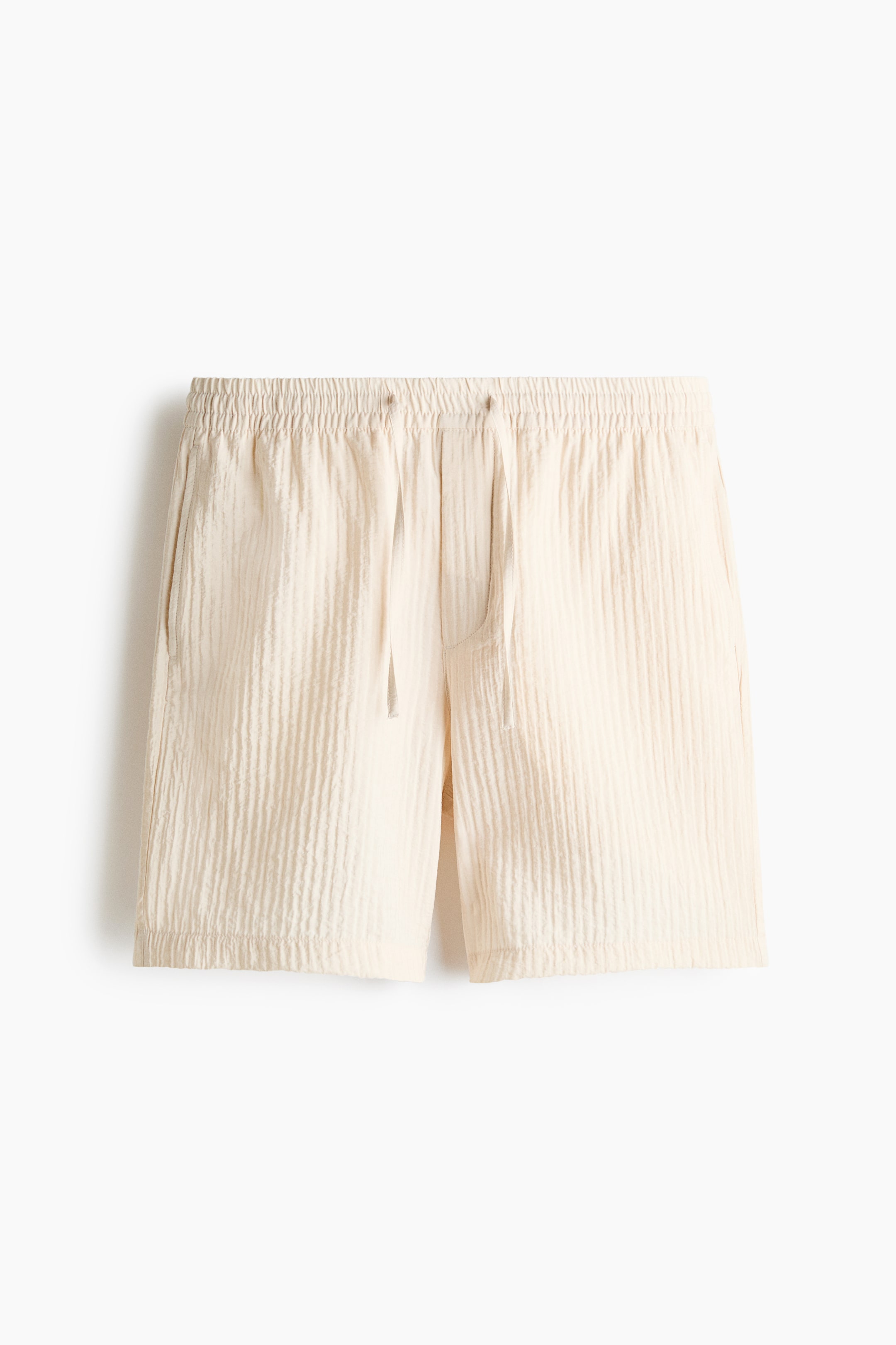 View larger image: The front of the shorts showcases a light beige hue with a vertical crinkled texture, featuring an elastic waistband with a drawstring and visible side pockets.