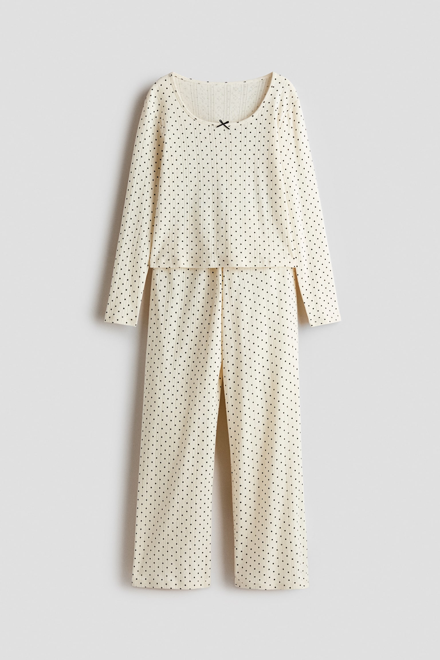 Ein zweiteiliges Pyjama-Set aus cremefarbenem Pointelle-Strick mit schwarzem Polka-Dot-Muster ist von vorne zu sehen. Das Set besteht aus einem Langarmoberteil mit Rundhalsausschnitt, verziert mit einer kleinen schwarzen Satinschleife, sowie einer passenden Hose mit geradem Bein, die darunter hÃ¤ngt.