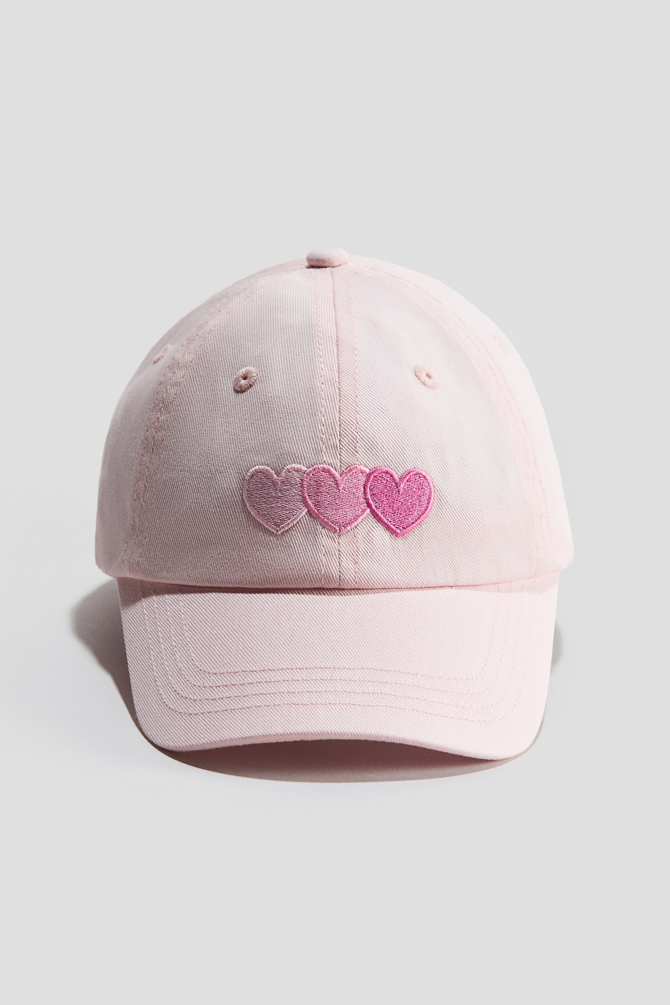 View larger image: "A pink cotton twill cap is displayed from the front view, showcasing three embroidered hearts arranged horizontally in different shades of pink on its front panel."