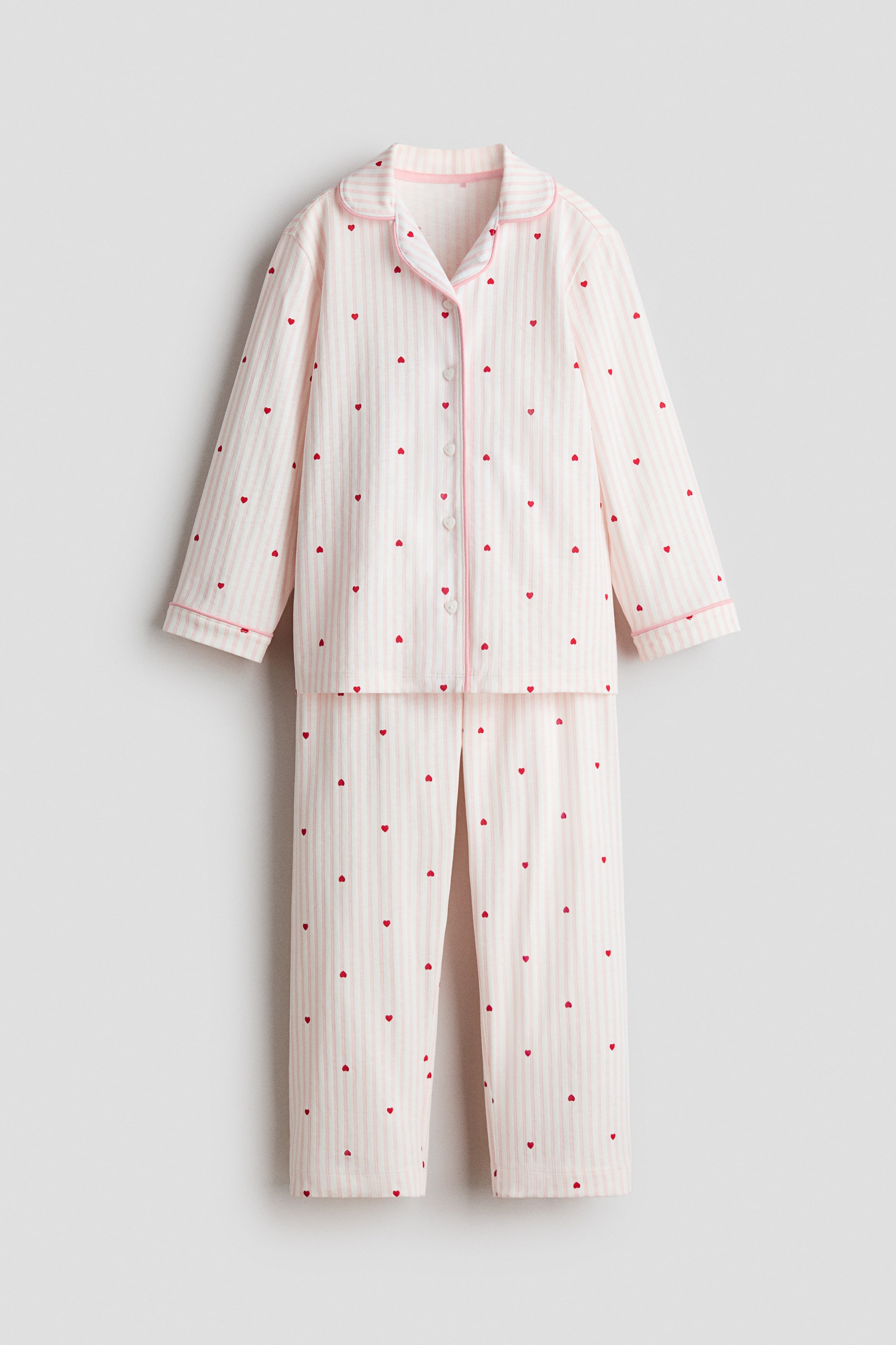 Ver imagen más grande: Un conjunto de pijama presenta rayas verticales en tonos rosa claro y blanco, acentuado con pequeños corazones rojos dispersos por todo el diseño. La parte superior de manga larga presenta un clásico cuello con muescas y ribetes rosados, un cierre frontal completo con botones en forma de corazón y ribetes rosados en los puños. Los pantalones de pierna recta a juego completan el conjunto.