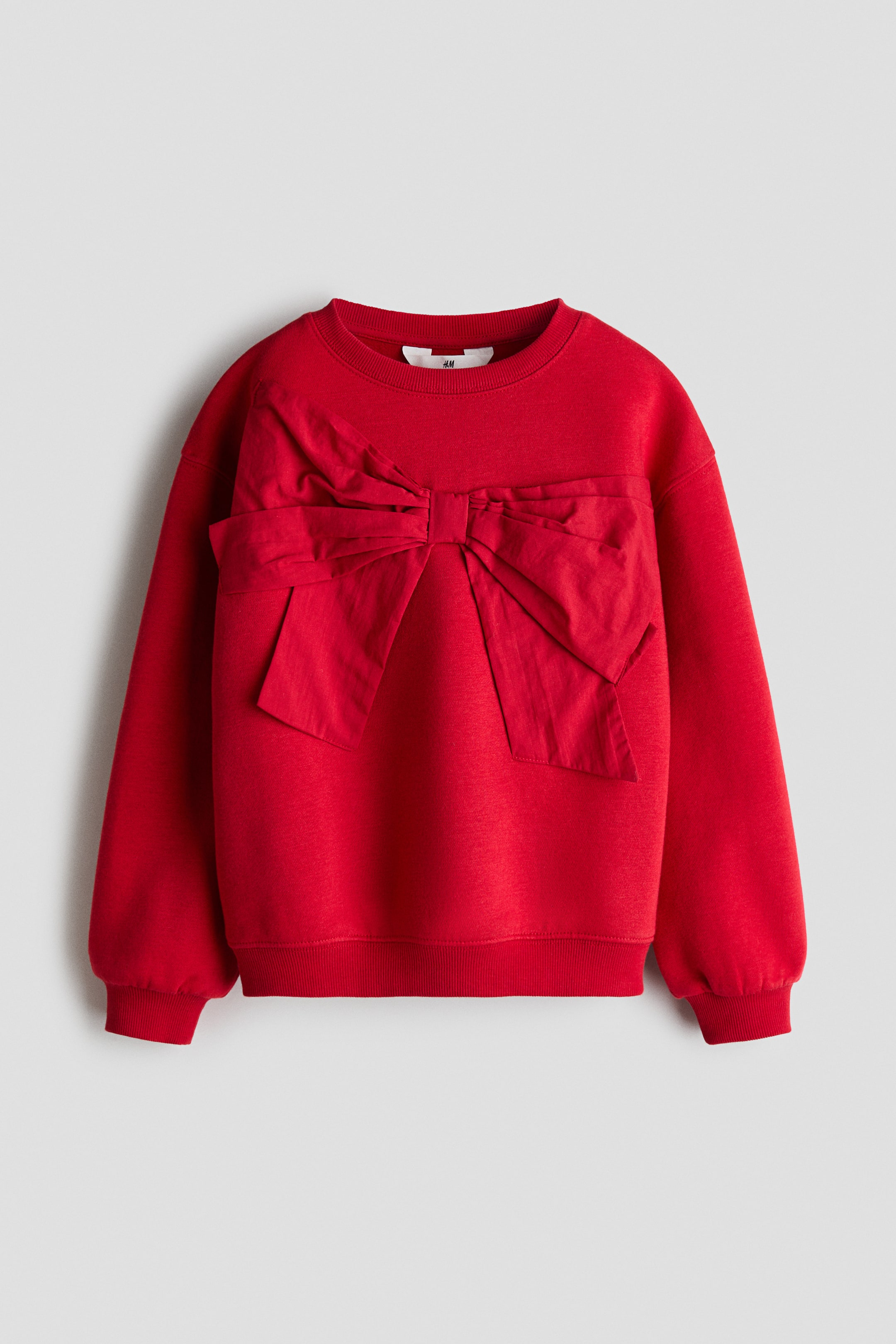 View larger image: A red sweatshirt is shown from the front, featuring a large decorative fabric bow stitched across the chest. The garment features a ribbed crew neck, dropped shoulders, ribbed cuffs, and a ribbed hem.