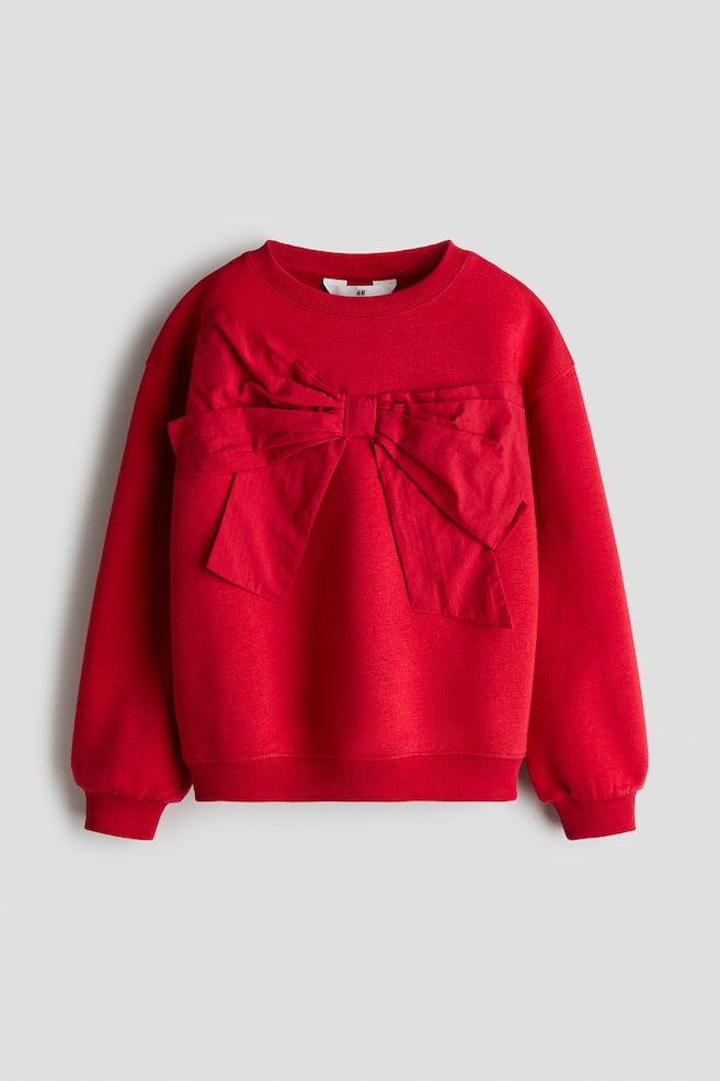 A red sweatshirt is shown from the front, featuring a large decorative fabric bow stitched across the chest. The garment features a ribbed crew neck, dropped shoulders, ribbed cuffs, and a ribbed hem. - 1