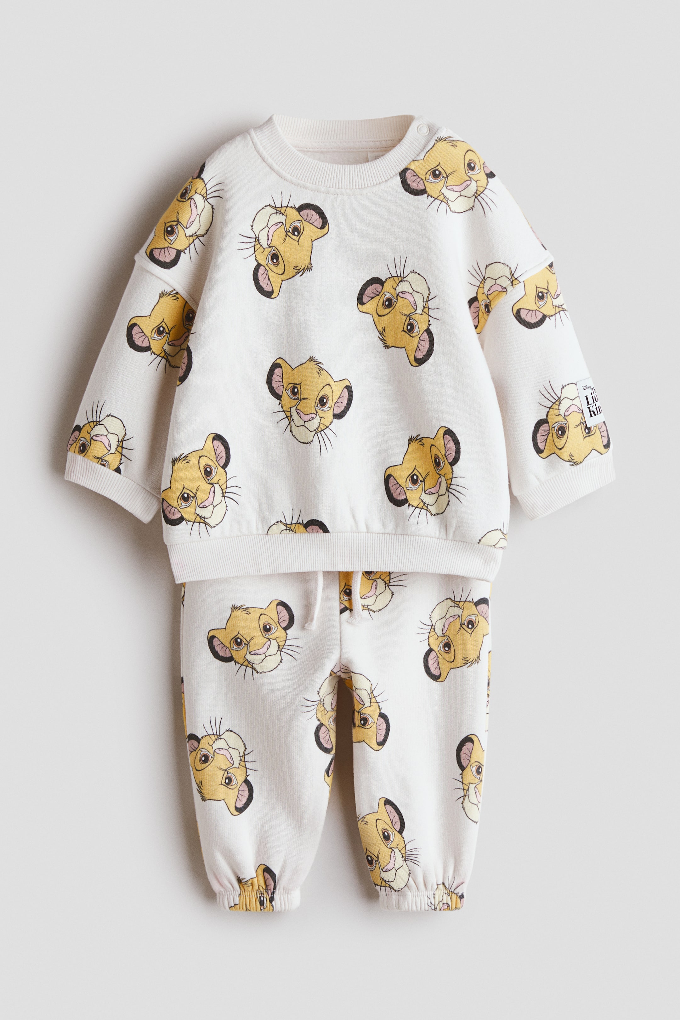 View larger image: A cream-coloured two-piece baby outfit is showcasing its front, featuring a long-sleeve sweatshirt and matching track pants. The entire ensemble displays an all-over print of cartoon lion cub faces. The sweatshirt boasts a crew neck, snap button closures on the shoulder, and ribbed cuffs and hem, while the track pants feature an elasticised waistband with a drawstring and ribbed ankle cuffs.