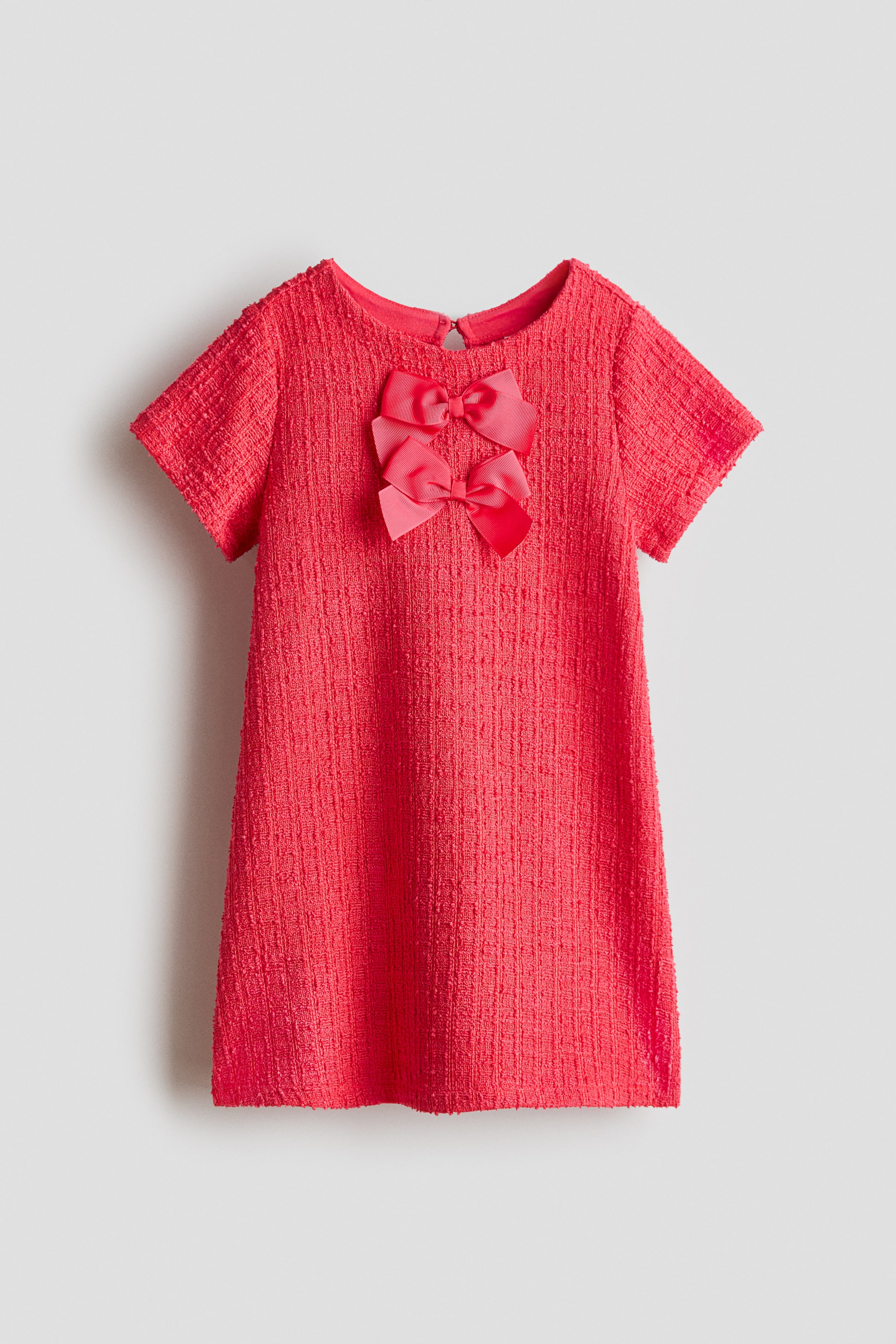 查看大圖: 這件短袖珊瑚色連身裙展現細緻織紋與圓領設計。胸前飾有兩個上下疊置的粉紅色蝴蝶結，後領處可見一個小型鑰匙孔開口。