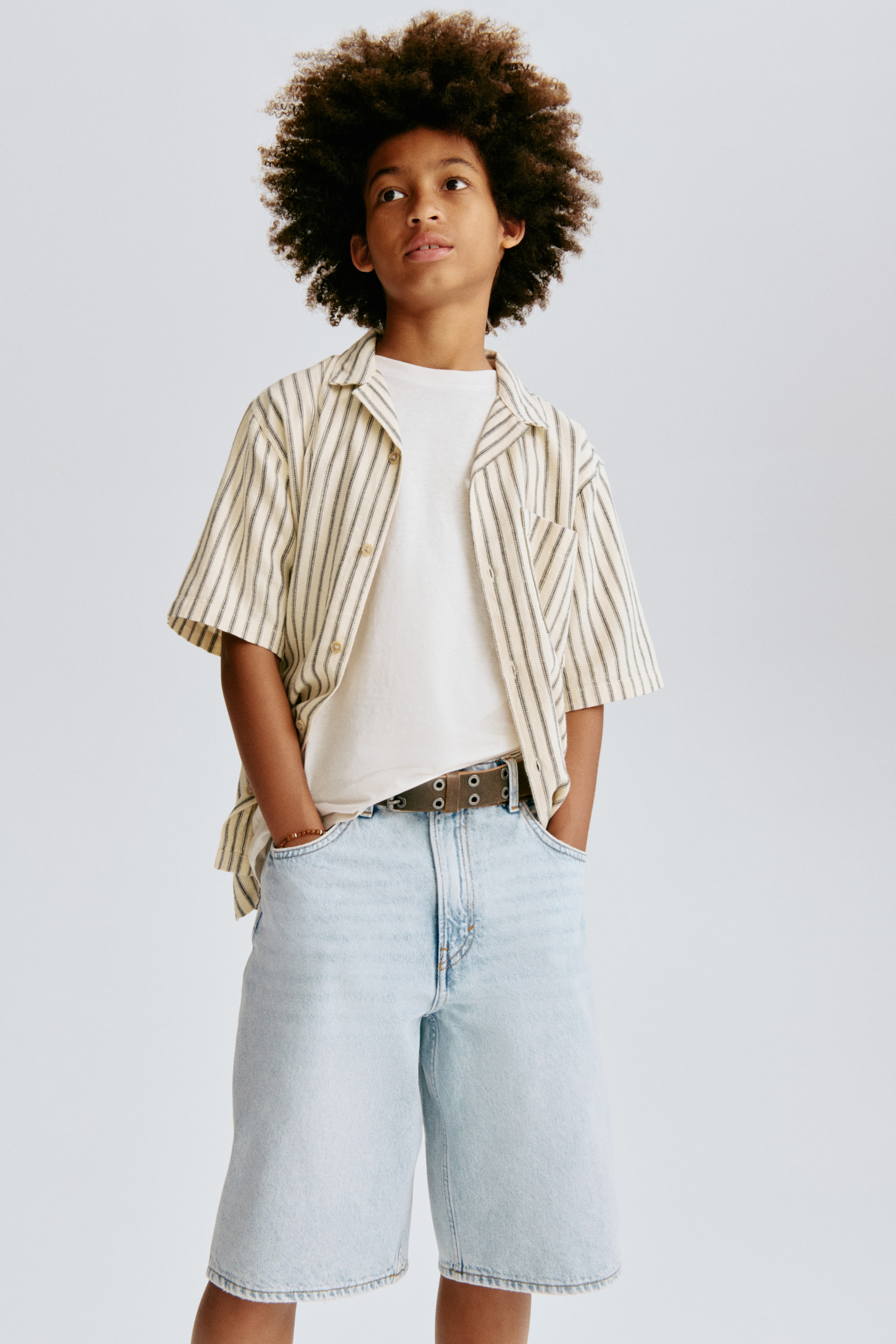 View larger image: Knee-length, light blue washed cotton denim shorts are depicted from the front, showcasing a relaxed fit through the hip and thigh with a dropped crotch. They feature front pockets and a zip fly with a button fastening, with a brown belt threaded through the belt loops.