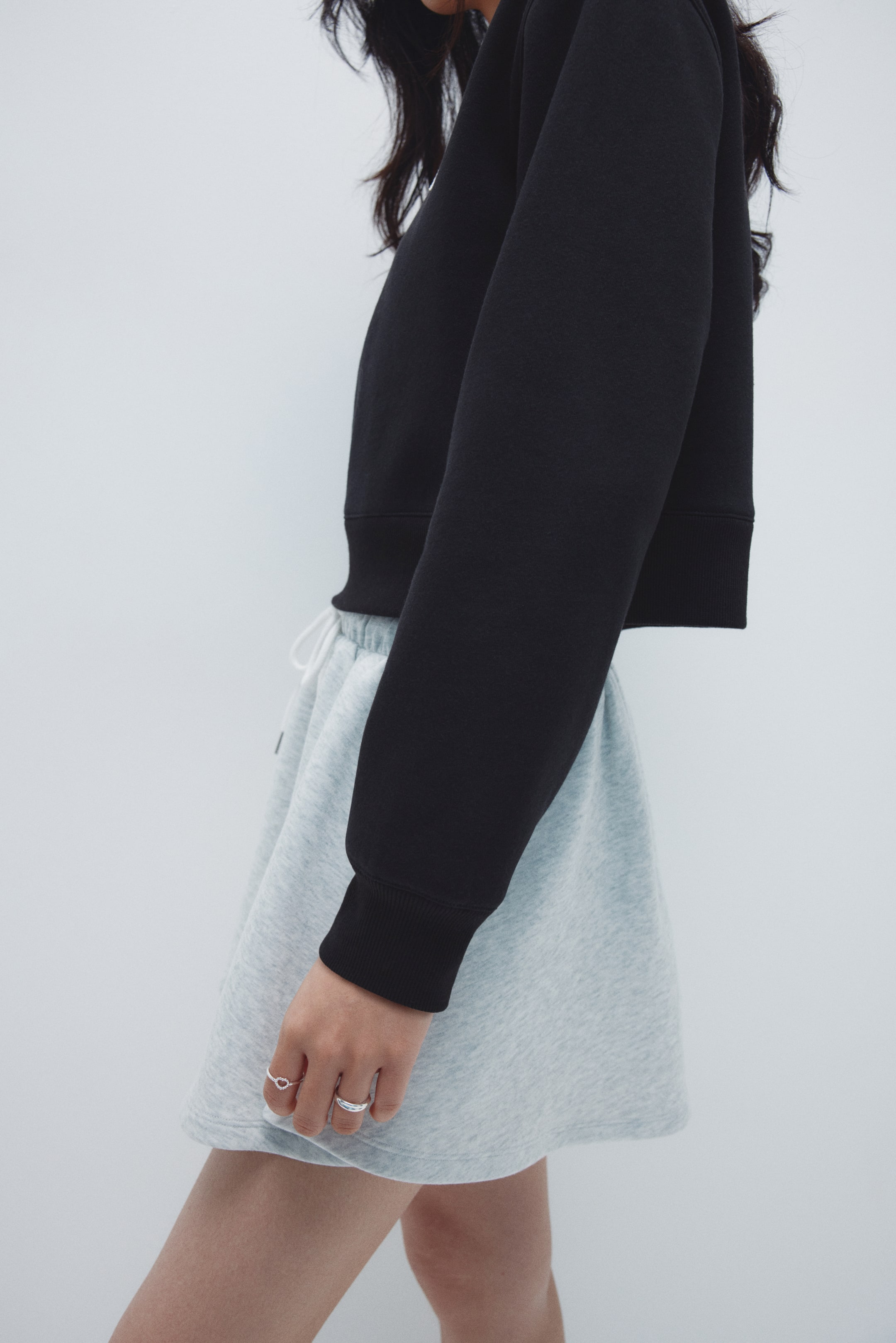 Xem ảnh lớn hơn: Một áo crop tay dài màu đen đang trình bày cổ tay và gấu áo có gân, được giới thiệu từ góc nhìn bên cạnh. Nó đang được mặc qua quần short xám nhạt có dây rút trắng ở eo.