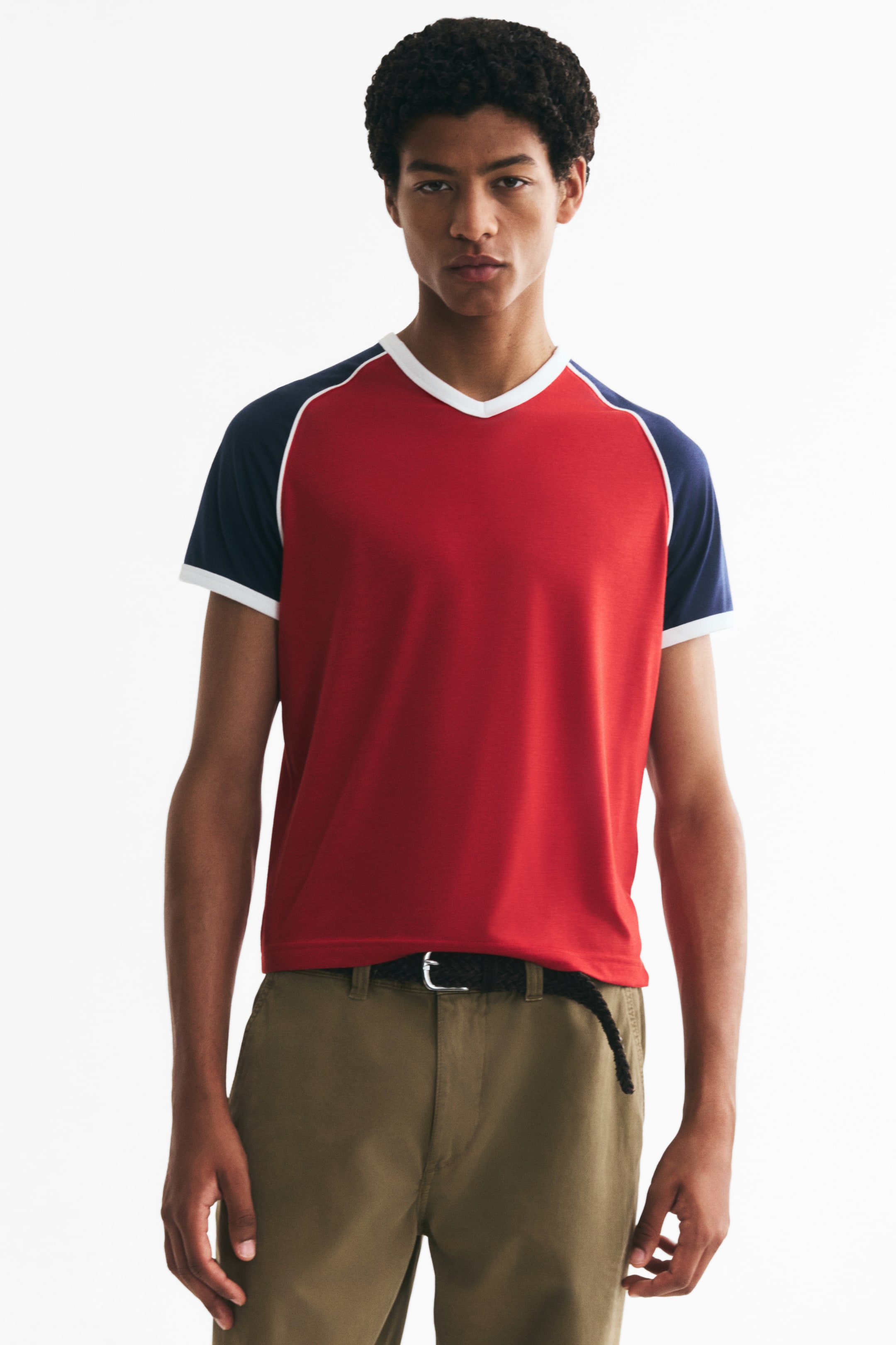 Visa större bild: En röd jersey-T-shirt med marinblå raglanärmar och vita kantningar vid V-ringningen och ärmsluten bärs av modellen. Den sitter slankt mot kroppen och är instoppad i olivfärgade byxor.