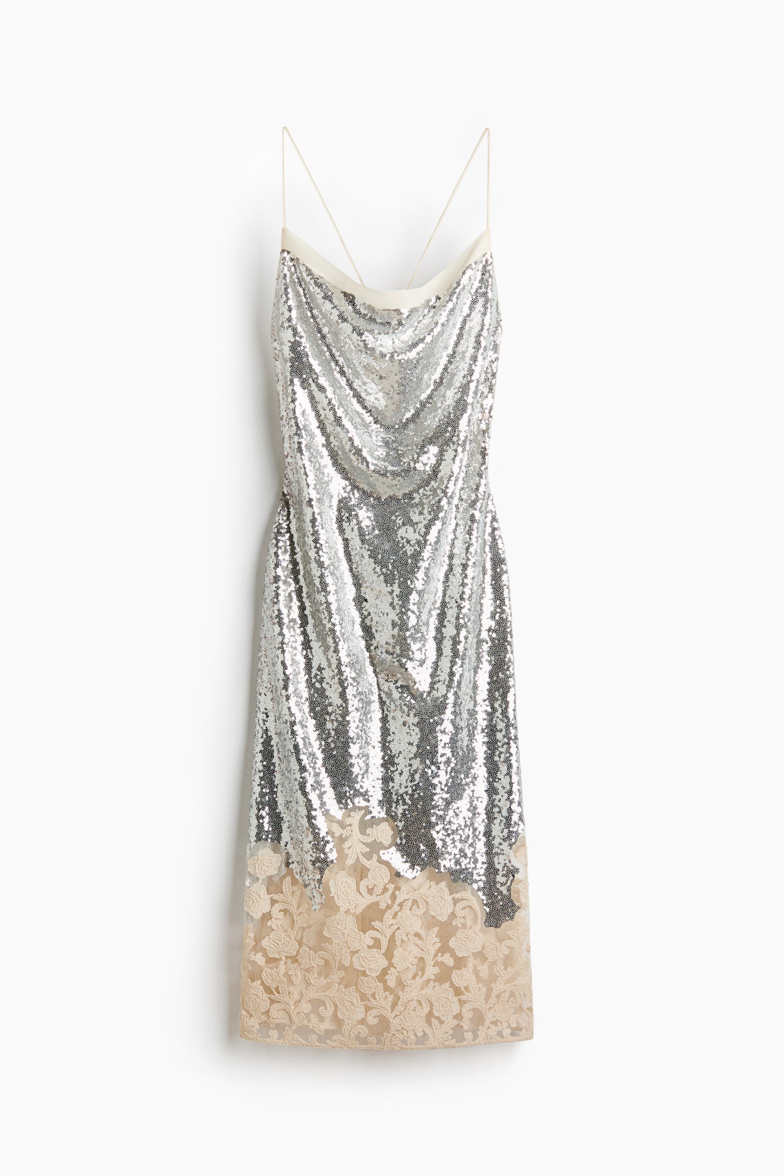 A silver sequinned slip dress is hanging, featuring a cream-coloured band above a draped cowl neckline and thin crossed straps. The bottom third of the dress is showcasing an intricate beige lace panel with a floral and scroll pattern. - 1