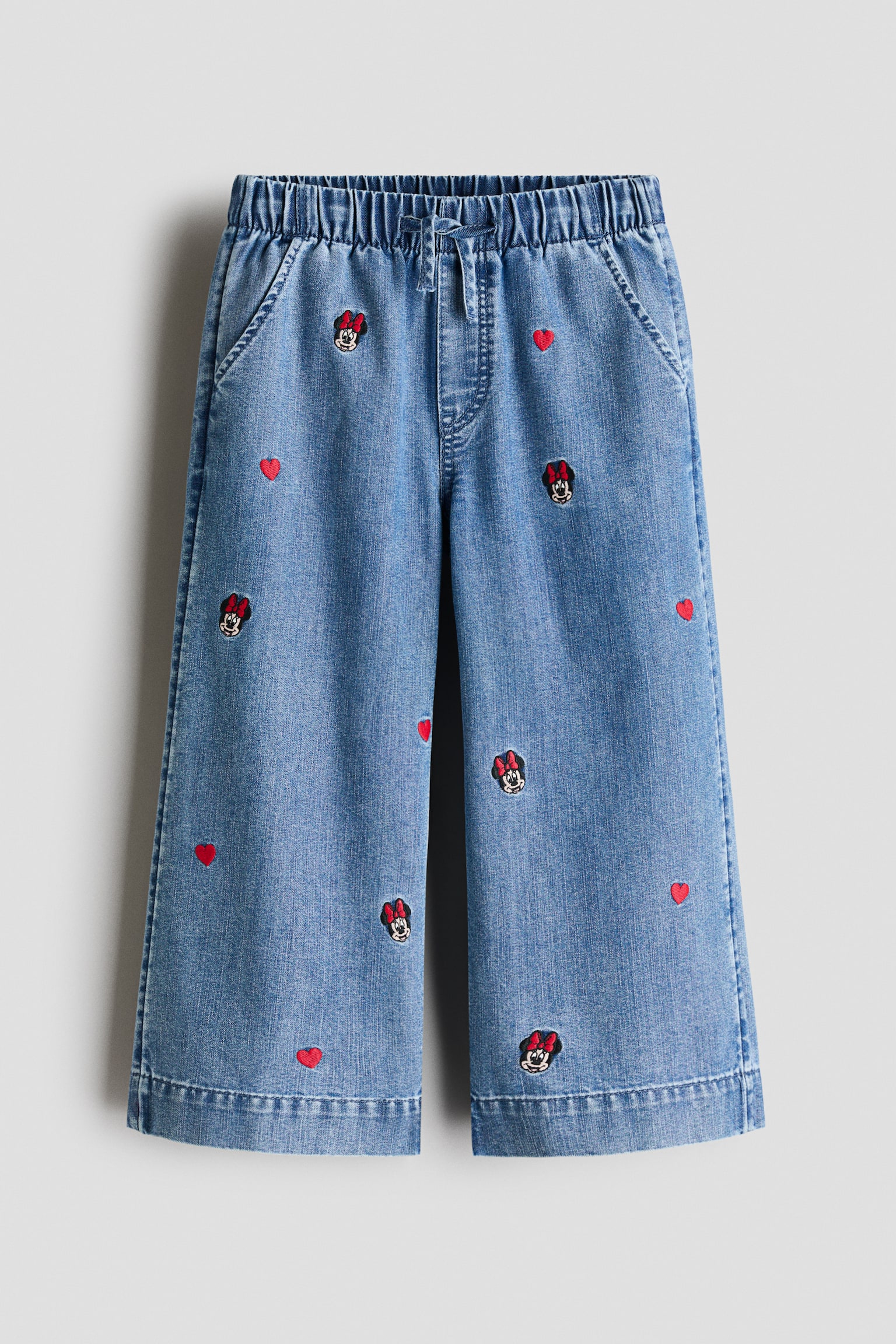 A pair of wide-leg denim joggers is shown from the front, featuring an elasticated waistband with a drawstring, a faux fly, and diagonal side pockets. The light blue denim is decorated with scattered embroidered Minnie Mouse motifs and small red hearts.