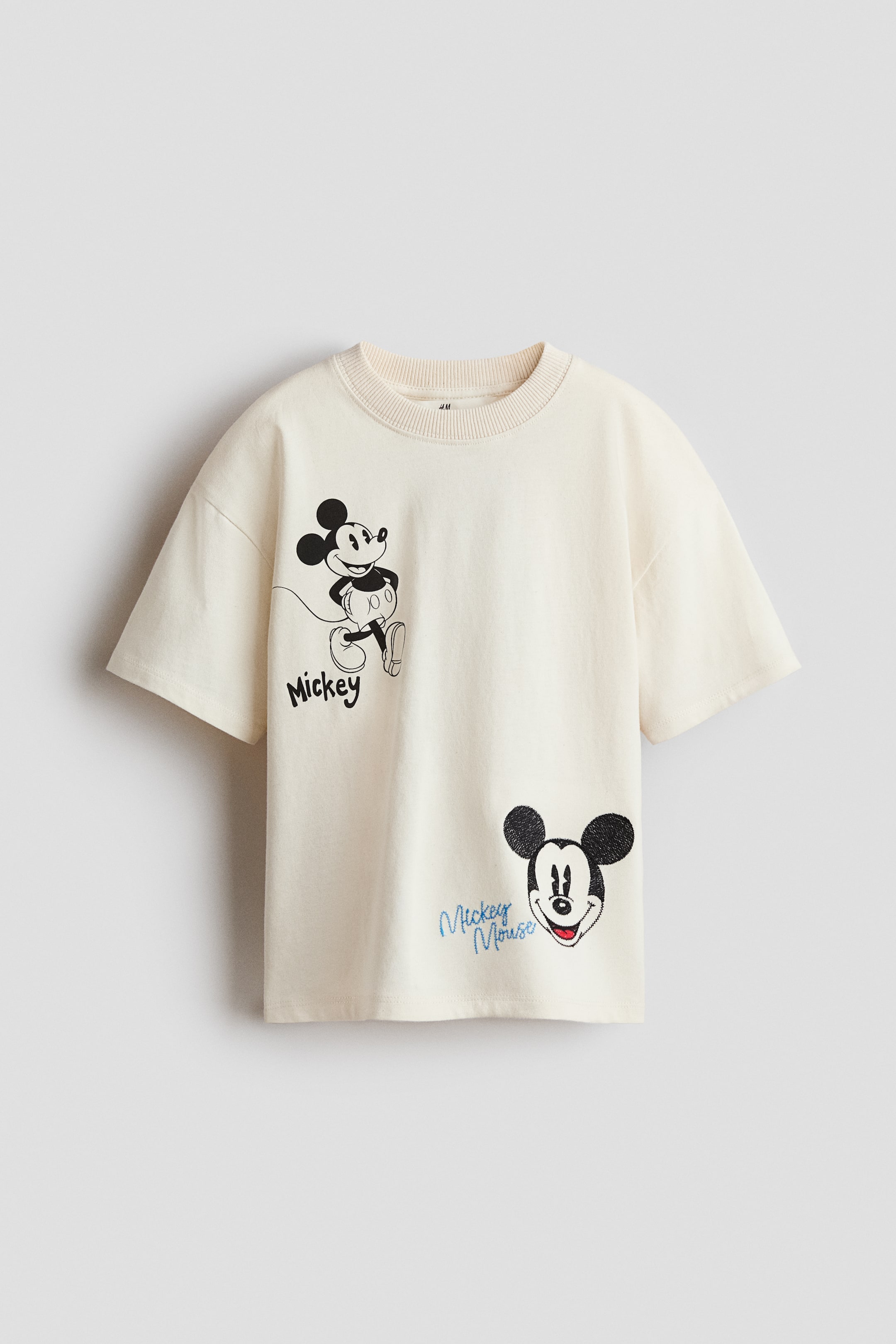 View larger image: A loose-fit, off-white, soft jersey t-shirt is shown facing front. It features a round, rib-trimmed neckline and dropped shoulders, with two black and white cartoon character print motifs: a full-body figure on the left with "Mickey" text, and a head on the lower right with "Mickey Mouse" text.