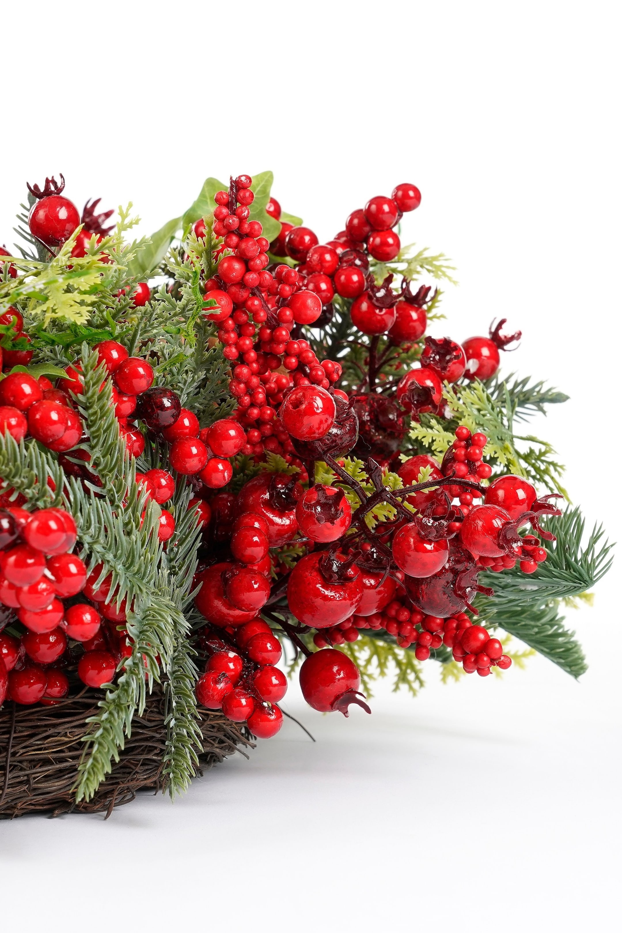 Grotere afbeelding bekijken: Een decoratieve opstelling toont een dichte groep glanzende rode bessen en groen loof, gepresenteerd vanuit een verhoogd, close-up perspectief. Verschillende maten van helderrode bessen vermengen zich met donkergroene dennennaalden en lichtere groene bladeren, die tevoorschijn komen uit een donkerbruine gevlochten basis.