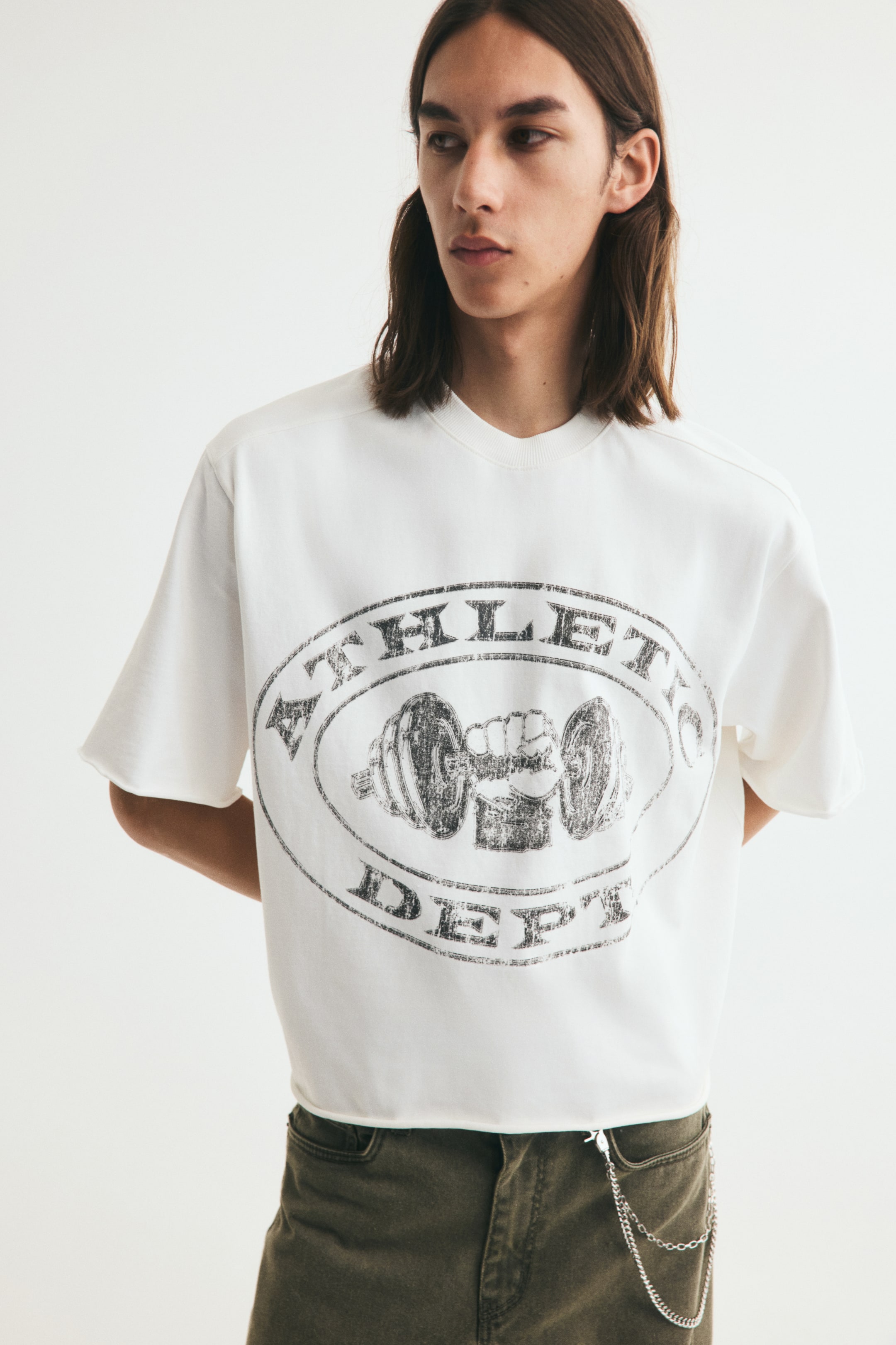 View larger image: A white, boxy-fit T-shirt featuring a distressed circular graphic with a dumbbell illustration and the words "ATHLETIC DEPT." is worn by the model. The T-shirt has a round neckline, dropped shoulders, and raw-cut edges, and is paired with olive-green trousers.