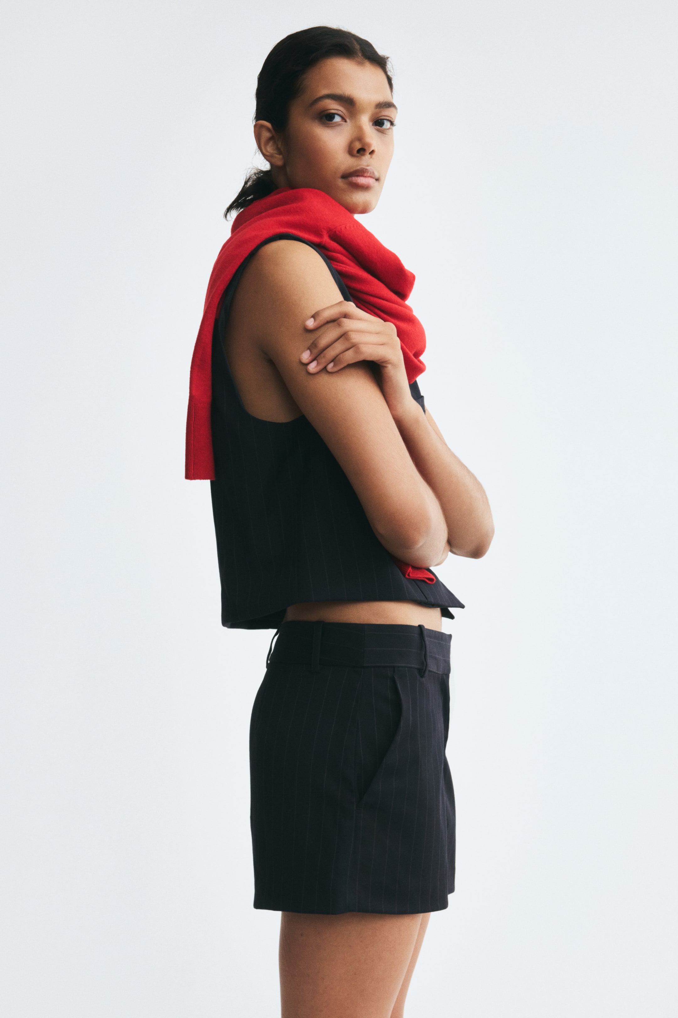 View larger image: A black pinstripe skort featuring a regular waist with belt loops and a diagonal side pocket is worn by the model. The skort is styled with a matching sleeveless pinstripe top and a bright red sweater draped over the shoulders.