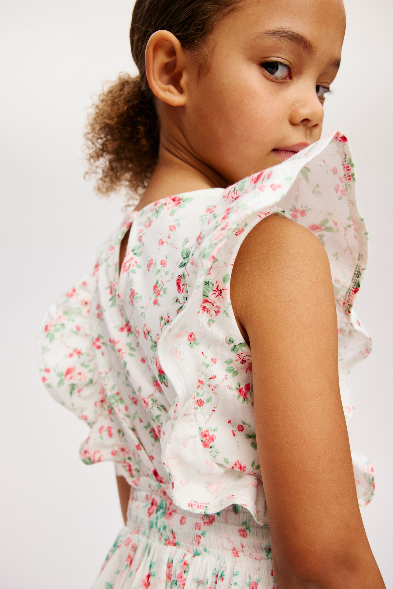 A white top is facing front, showcasing a floral pattern of pink roses and green leaves. It features short, ruffled cap sleeves, a round neckline, and vertical ruffles on the front bodice, with a keyhole closure and button visible at the back of the neck. - 1