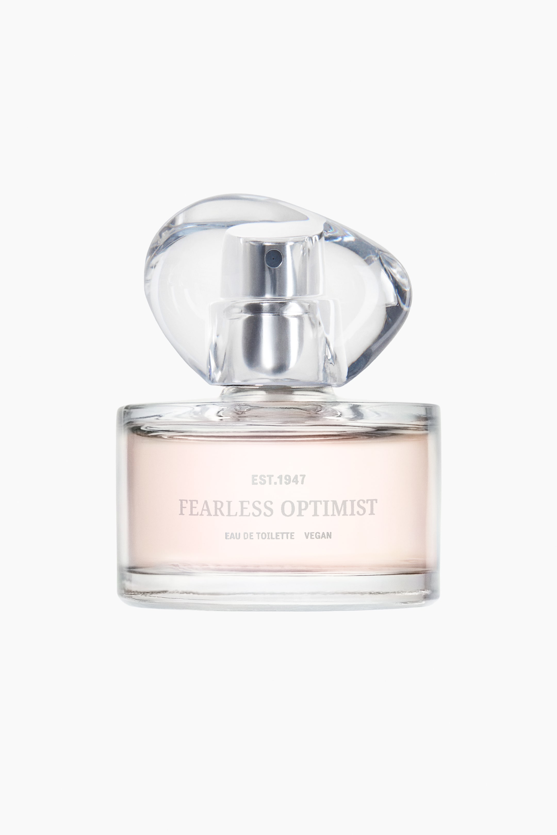 הצגת תמונה גדולה יותר: הבושם "The Fearless Optimist EdT" מוצג בקדמת הבמה, עם בקבוק זכוכית מלבני שקוף המלא בנוזל ורדרד בהיר. פקק זכוכית שקוף בצורת לא סדירה מכסה את הבקבוק, חושף מתז בגוון כסף, כאשר הכיתוב "Fearless Optimist" ו-"Eau de Toilette Vegan" מופיעים בטקסט אפור בחזית.