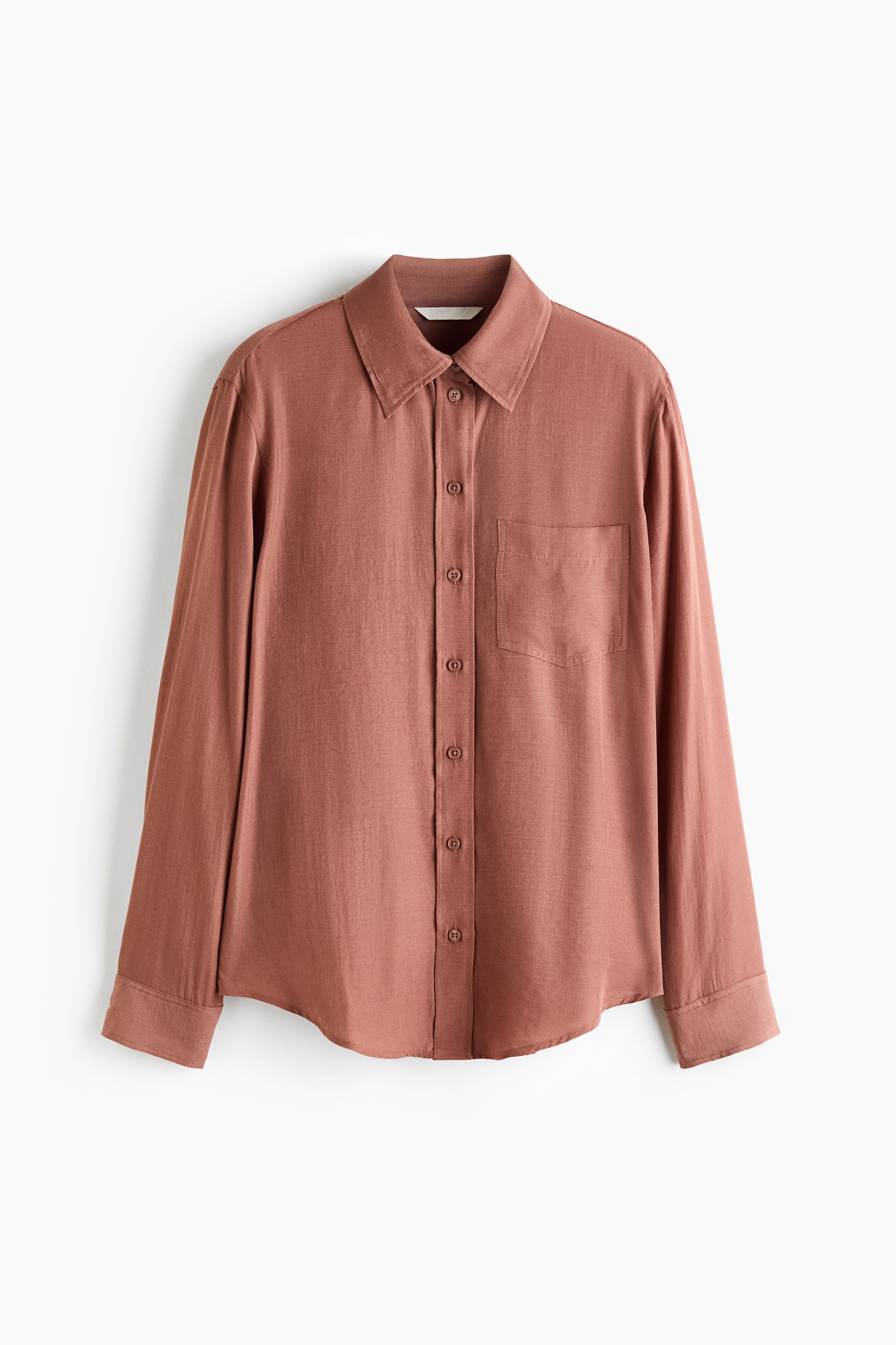 A terracotta-colored shirt in a soft weave is worn by the model with arms crossed. The shirt features a collar, a single patch chest pocket, long sleeves with button cuffs, and a rounded hem, styled with white trousers.