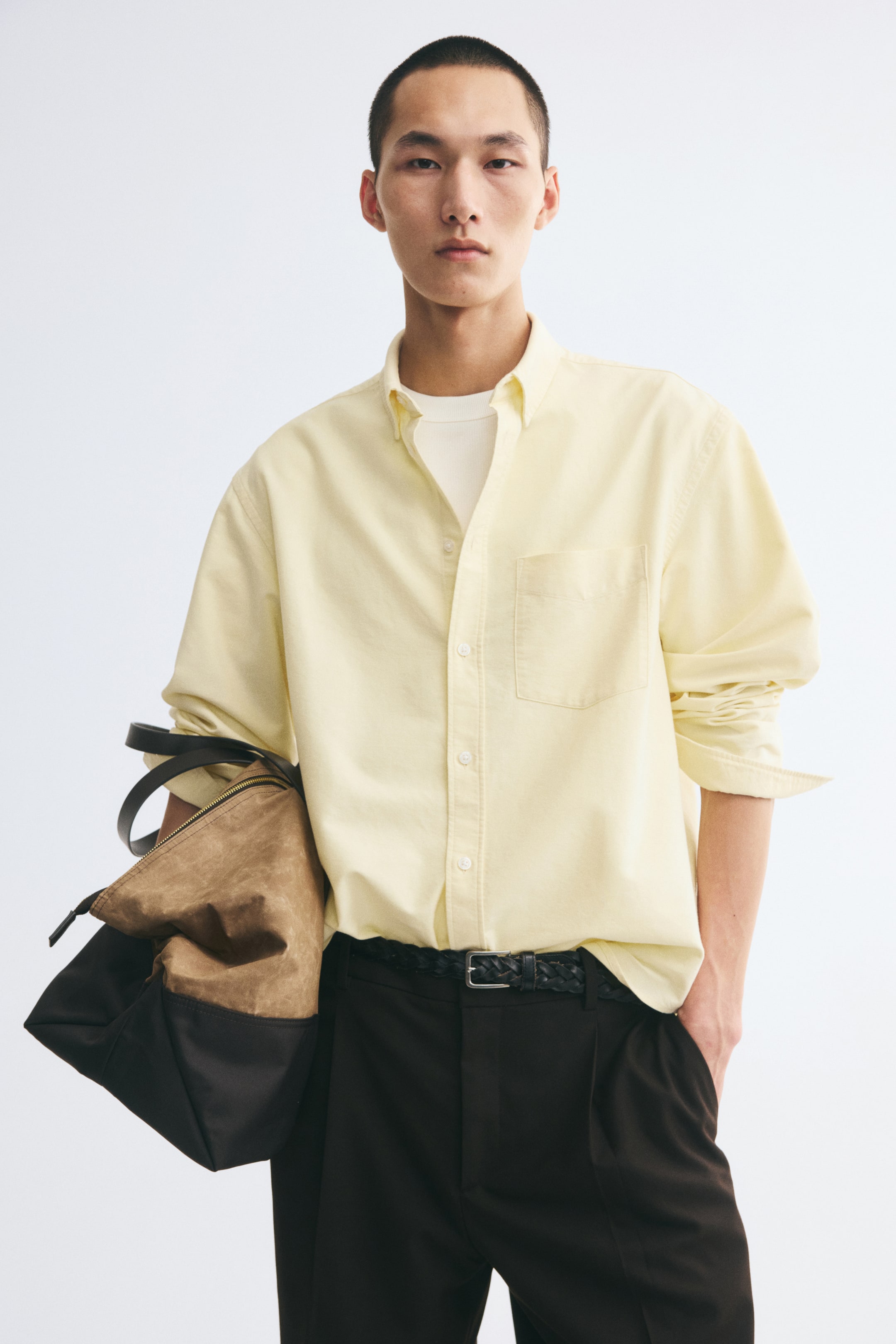 View larger image: The model is wearing a pale yellow Oxford cotton shirt layered over a white T-shirt and tucked into dark trousers. The shirt features a button-down collar, a classic button-front closure, a single chest pocket, and long sleeves rolled up at the cuffs.