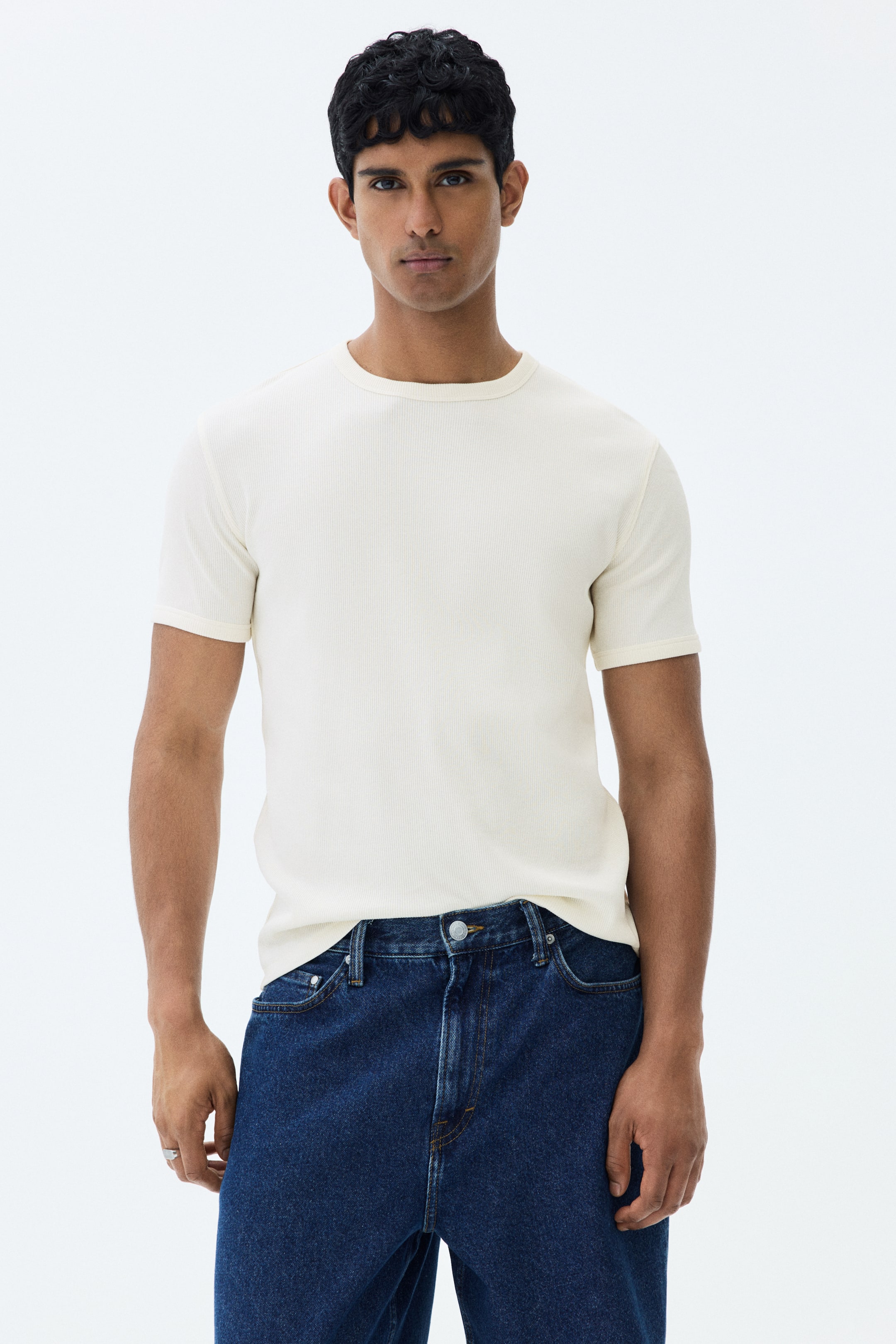 View larger image: The model is wearing a cream ribbed cotton jersey t-shirt with a round neckline and short sleeves. This slim-fit t-shirt is neatly tucked into dark blue denim jeans.