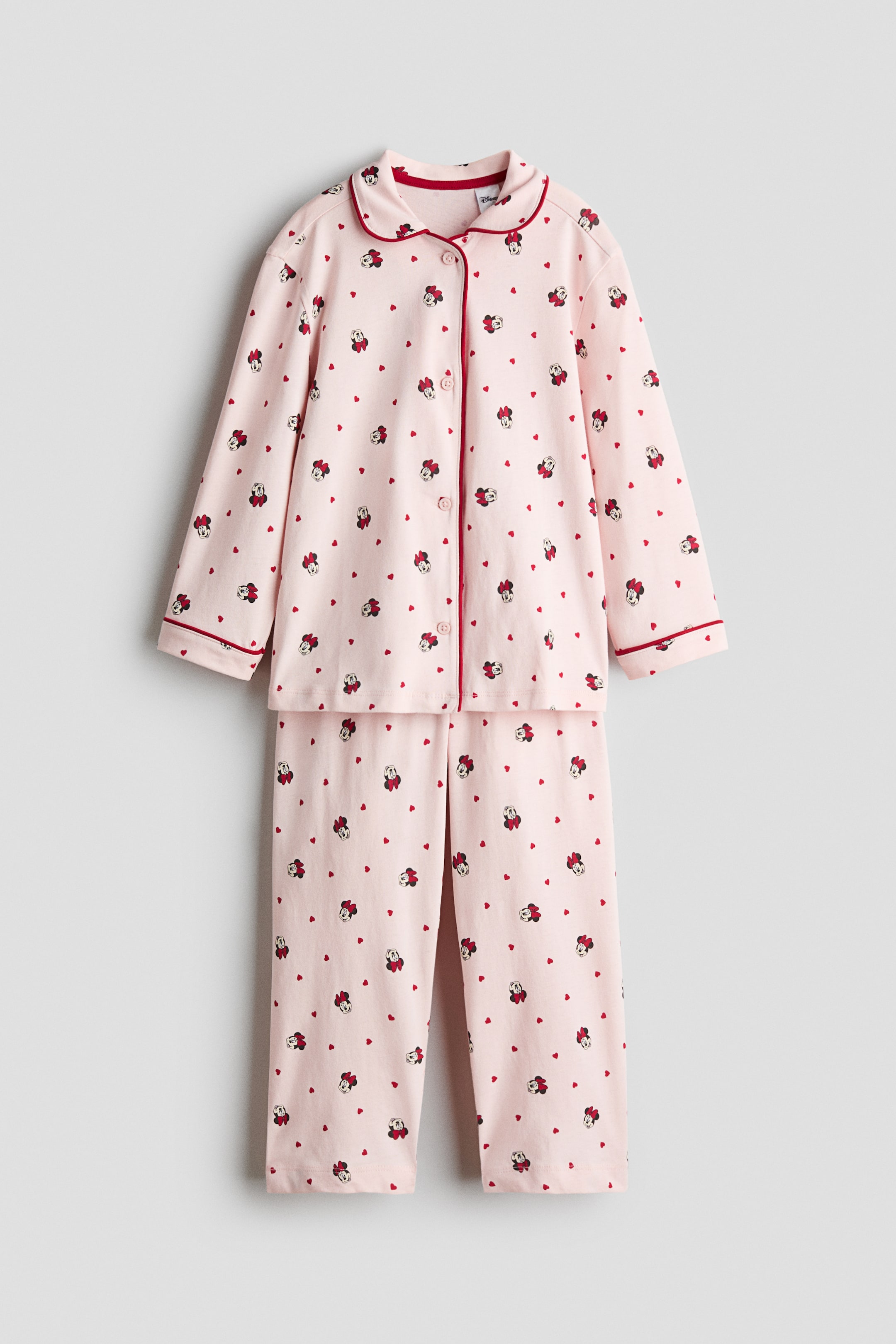 Größeres Bild anzeigen: Ein hellrosa Pyjama-Set in Frontansicht ist zu sehen, bestehend aus einem langärmeligen Hemd und einer passenden Hose, die mit einem Allover-Print aus Minnie Mouse und roten Herzen versehen sind. Das Hemd verfügt über einen runden Kragen, eine durchgehende Knopfleiste vorne sowie rote Kontrastpaspel entlang des Kragens, der Knopfleiste und der Ärmelbündchen.