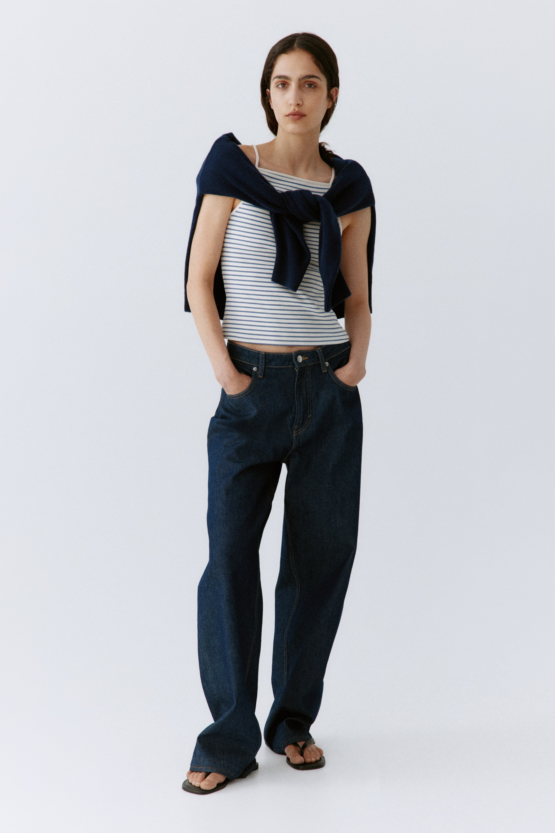 Visa större bild: En vit linne med tunna blå horisontella ränder, tunna axelband och en ribbad textur. Den bärs med mörktvättade, högmidjade, vida jeans som visar en synlig knapp- och dragkedjestängning. En marinblå tröja är knuten runt axlarna, och svarta platta tåsandaler syns under jeansen.
