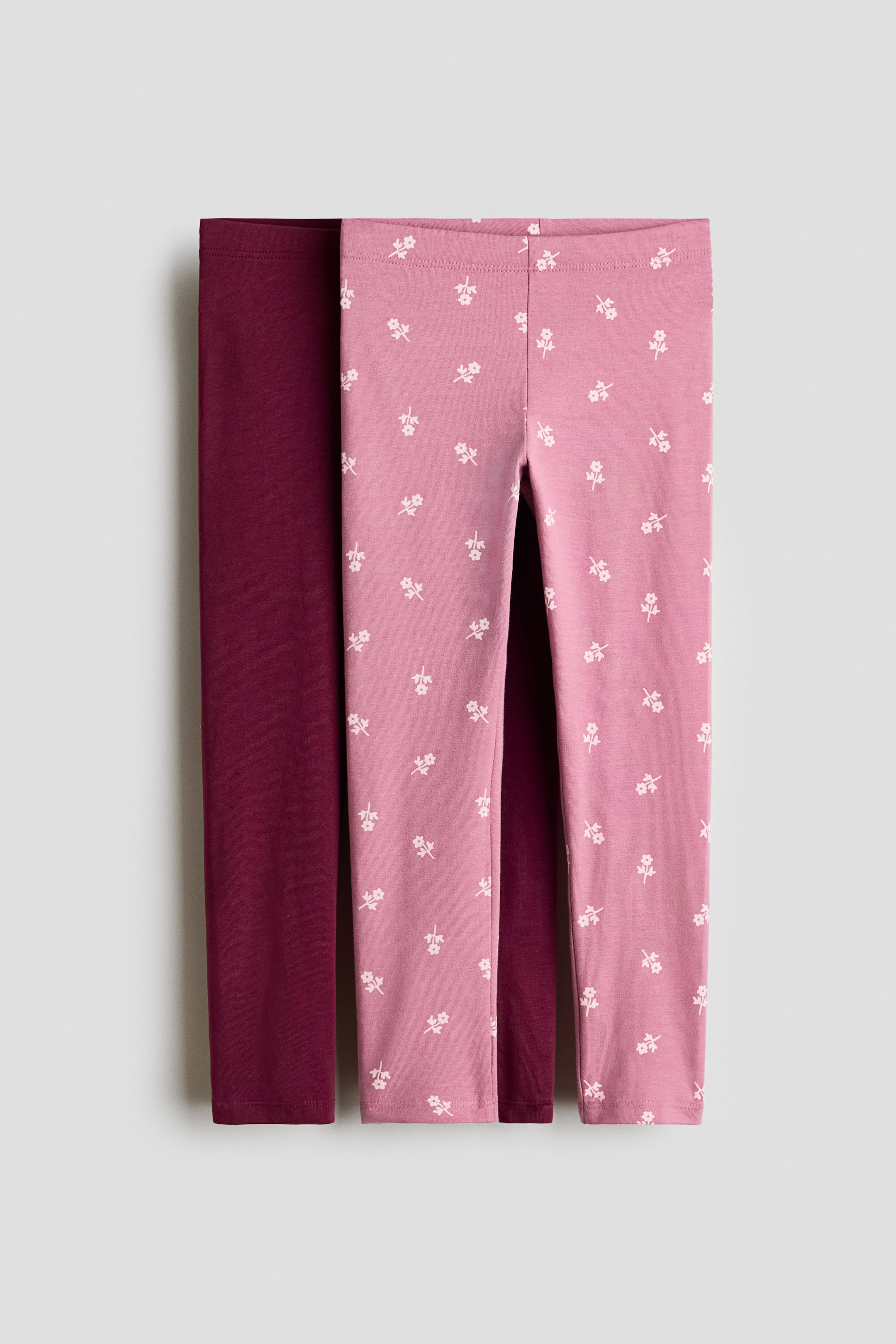 View larger image: Two pairs of jersey leggings are showcased, displaying their front view. One pair is a solid burgundy color, while the other is pink adorned with a repeating pattern of small white flowers. Both pairs come with an elasticated waist.