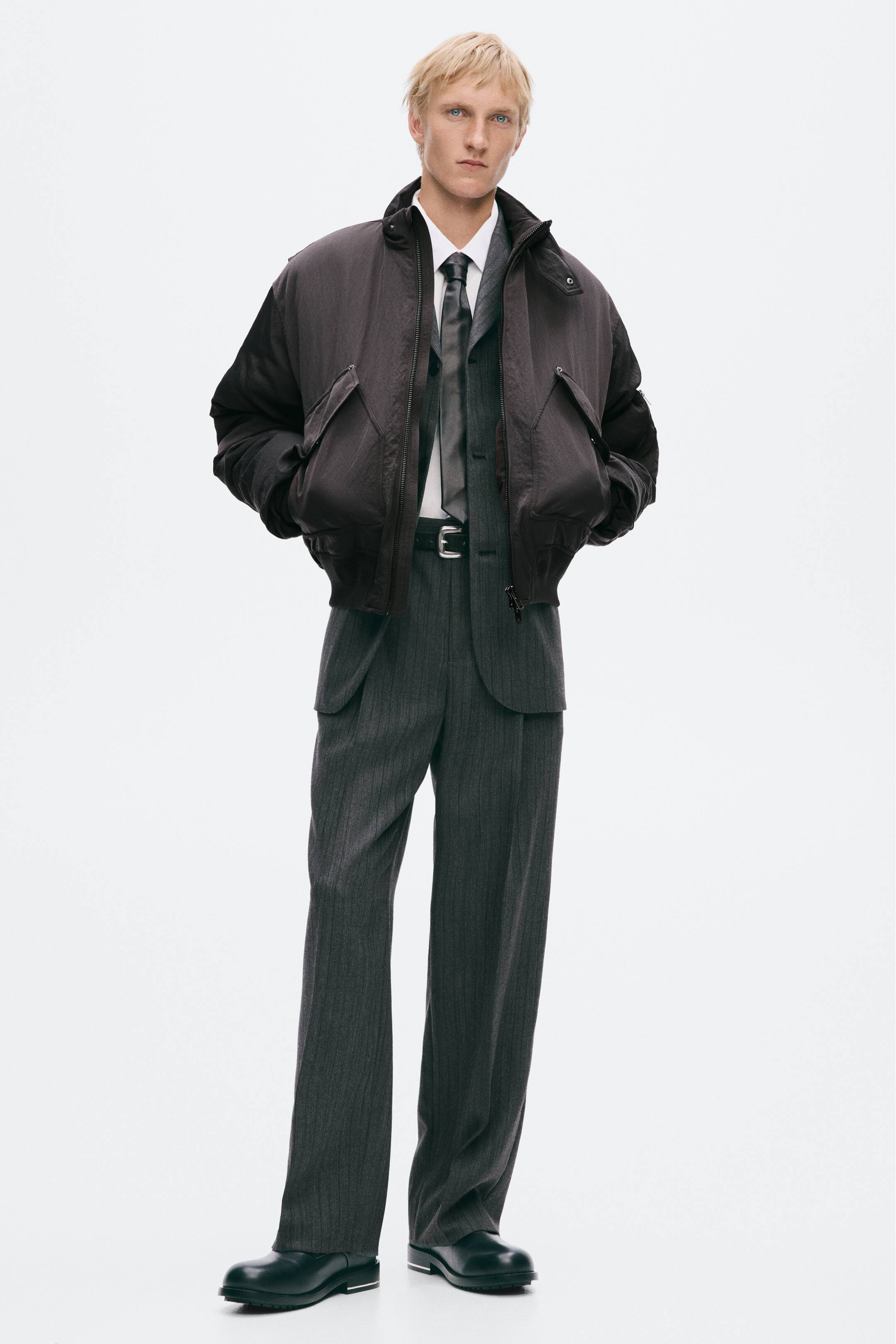 View larger image: A dark brown oversized padded bomber jacket with a high collar and large front flap pockets is being worn by the model, with their hands tucked into the pockets. The jacket is layered over a grey pinstripe suit, a white collared shirt, a grey tie, and black leather shoes.