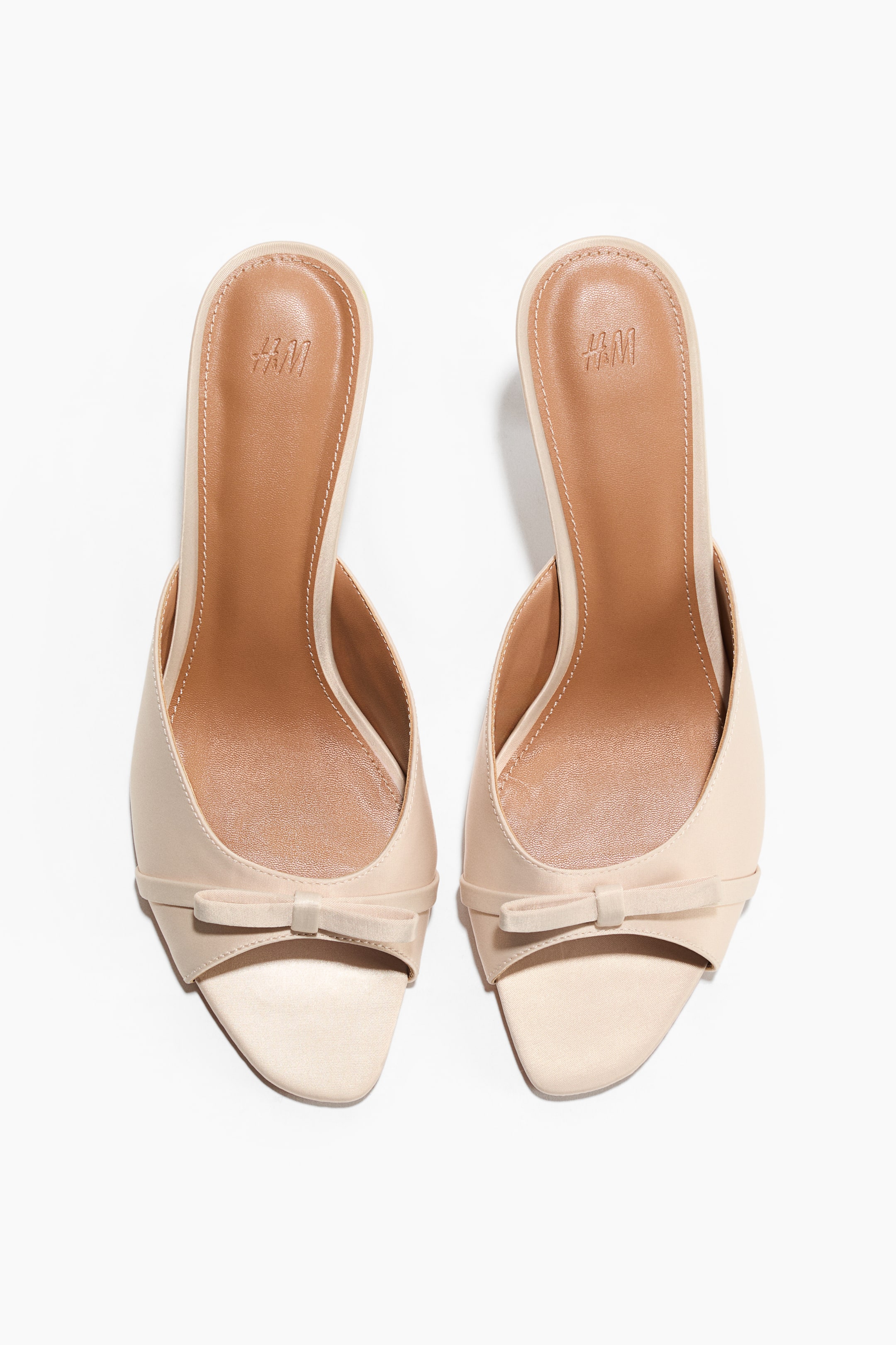 Visa större bild: Ett par ljusbeige mules presenteras från en något framåtvänd, uppifrån-och-ner-vy, med spetsiga tår och en liten rosett-detalj på vristen. Den bruna innersulan på varje mule visar en präglad "H&M"-logotyp.