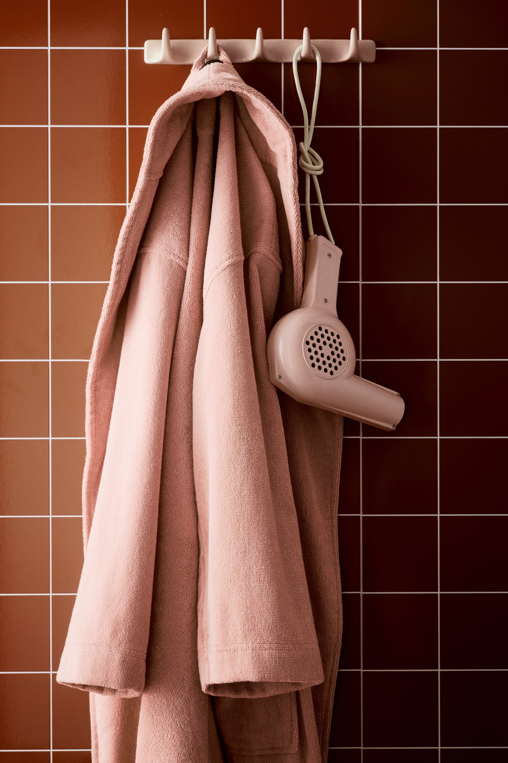 View larger image: A pink terry cloth bathrobe is hanging from a hook, its soft fabric draping downwards. The bathrobe features a visible hood and sleeves, showing its absorbent texture.