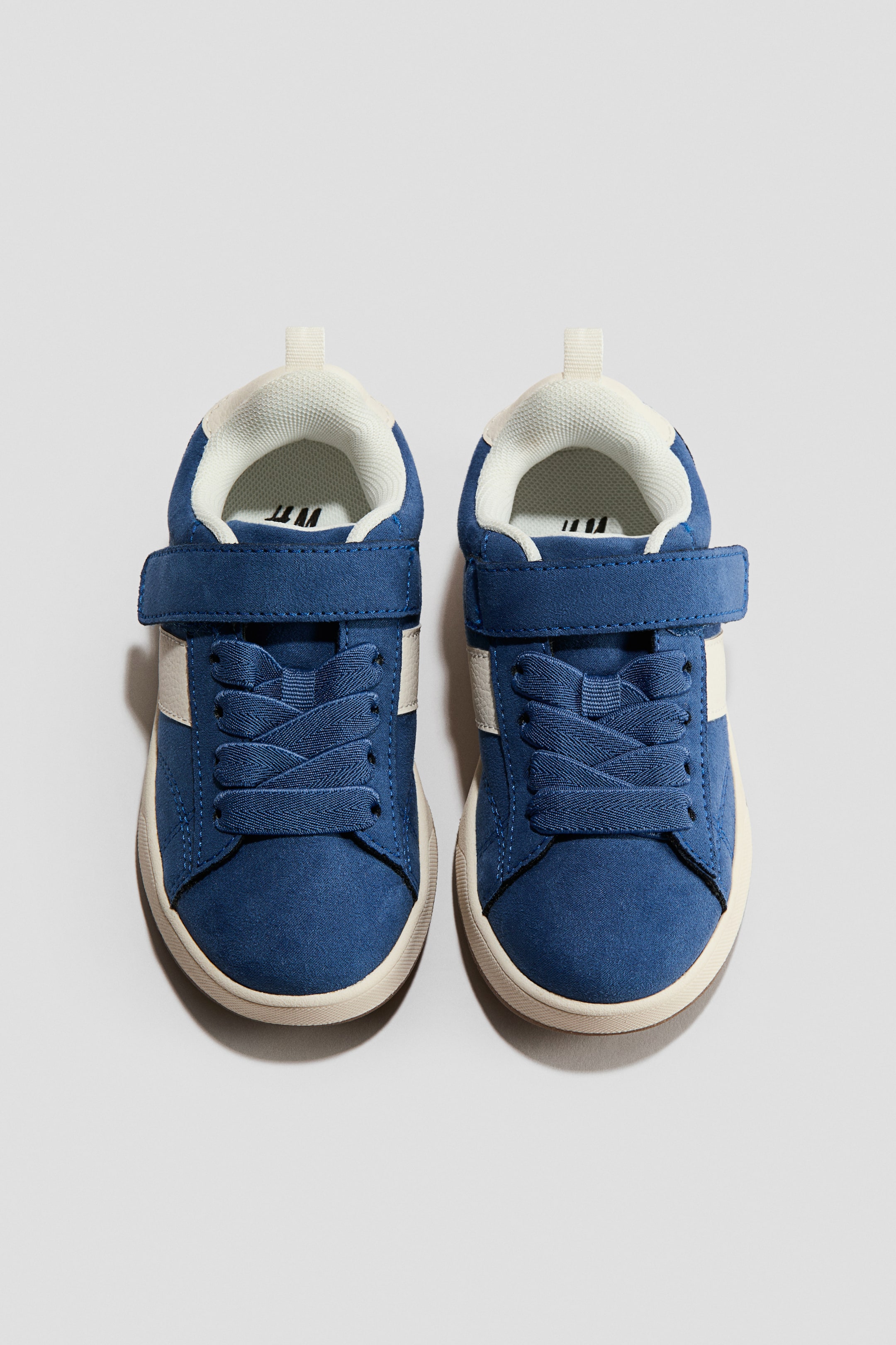 Ingrandisci l'immagine: Le scarpe da ginnastica sono riprese da un'angolazione frontale: un paio blu con tomaia a effetto scamosciato, suole in gomma color panna, collarini imbottiti e una striscia bianca sul fianco. Presentano lacci blu e una chiusura a strappo (hook-and-loop) sul collo del piede.