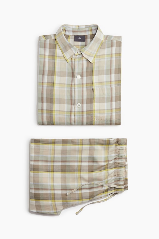 A folded pajama set is displaying a multi-toned plaid pattern in beige, brown, and yellow hues. The shirt is featuring a pointed collar, a button-down front with white buttons, and a single chest pocket, and the matching pants are showing an elasticized waistband with a drawstring. - 1