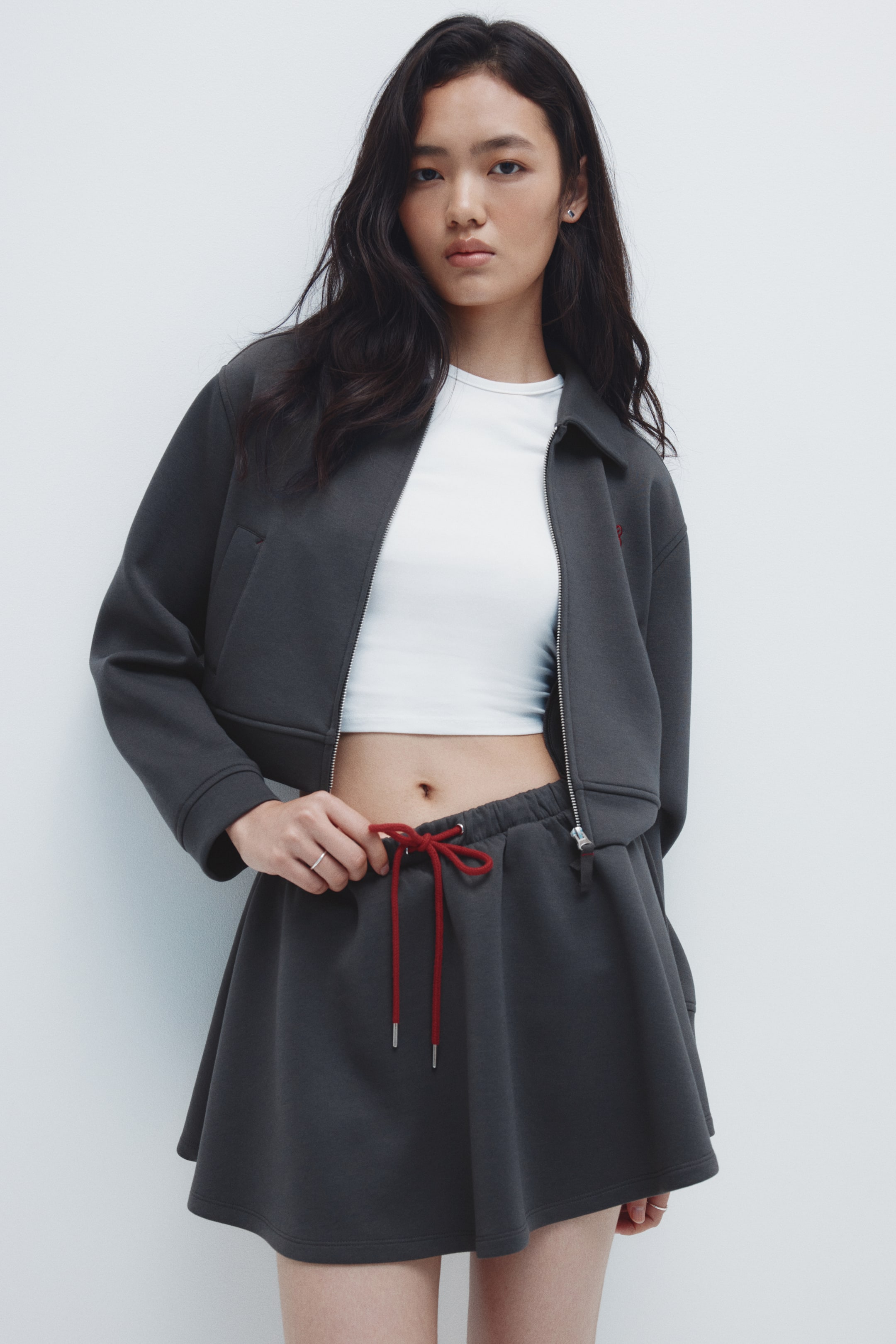 拡大画像を表示: ダークグレーのクロップドジャケットは、フルジップのフロント、尖った襟、フロントウェルトポケット、胸の小さな赤い刺繍が特徴で、白いクロップトップの上にオープンで着用されています。これに合わせて、同じダークグレーのフレアミニスカートがエラスティックウエストと赤いドローストリングが見えるデザインで登場しています。