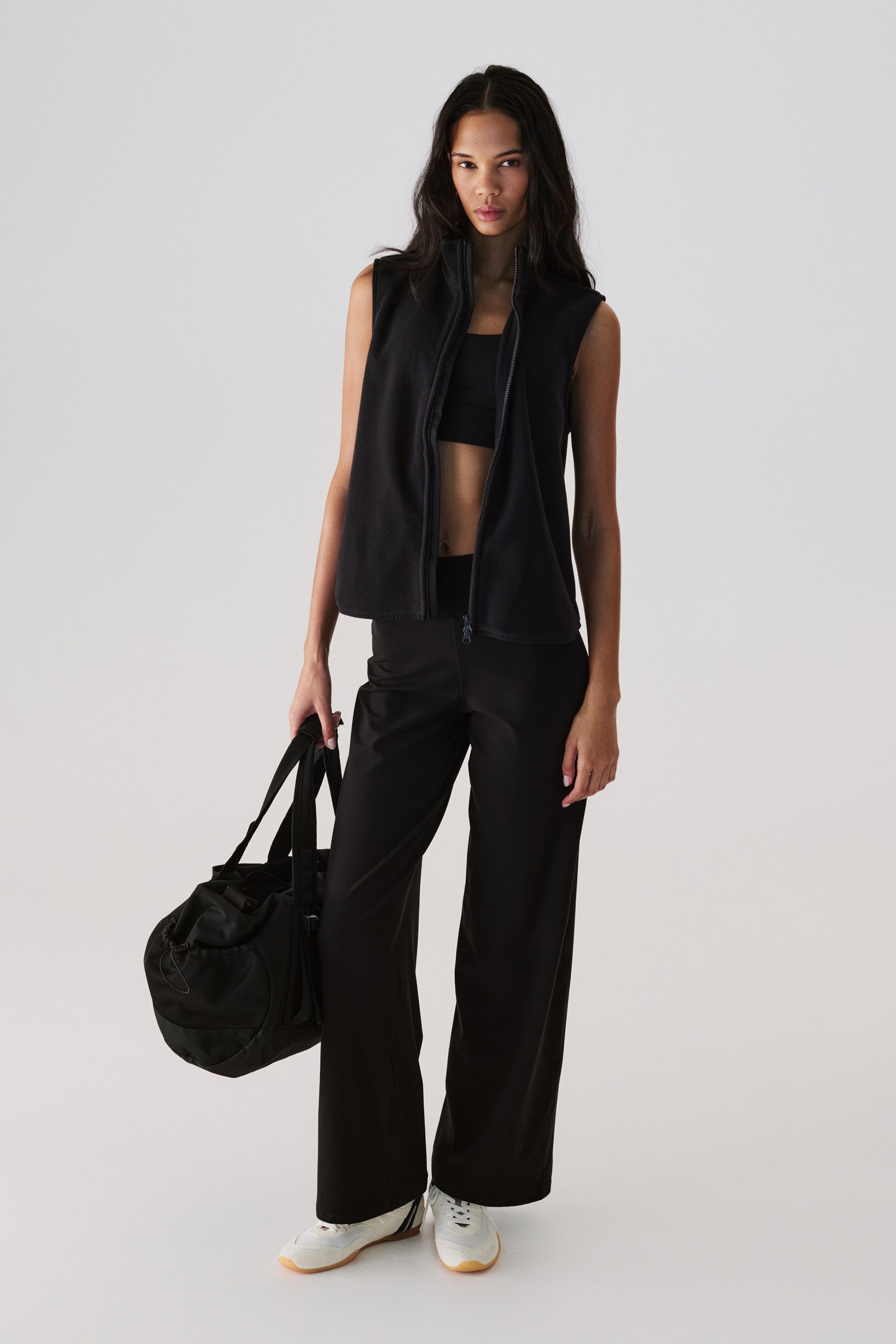 Ampliar la imagen: Se presenta un chaleco sin mangas de forro polar negro desde una vista frontal, con cierre de cremallera completa y cuello alto, y se lleva abierto sobre un sujetador deportivo negro.