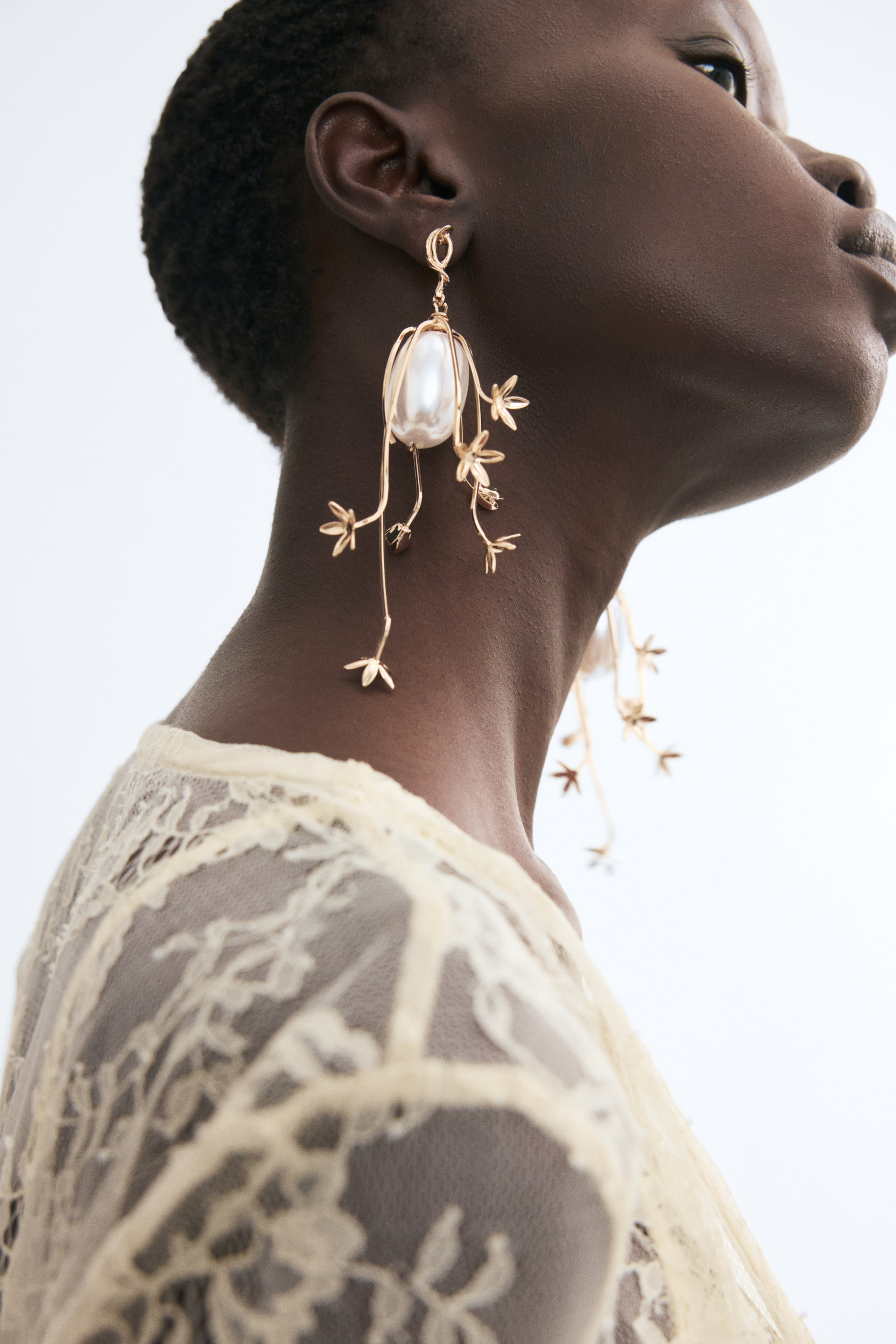 View larger image: Metal earrings featuring delicate, flower-shaped gold-toned pendants draped over a large pearlescent plastic bead are being worn by the model. The earrings hang from the earlobe, with thin, curved metal branches dangling down towards the neck.