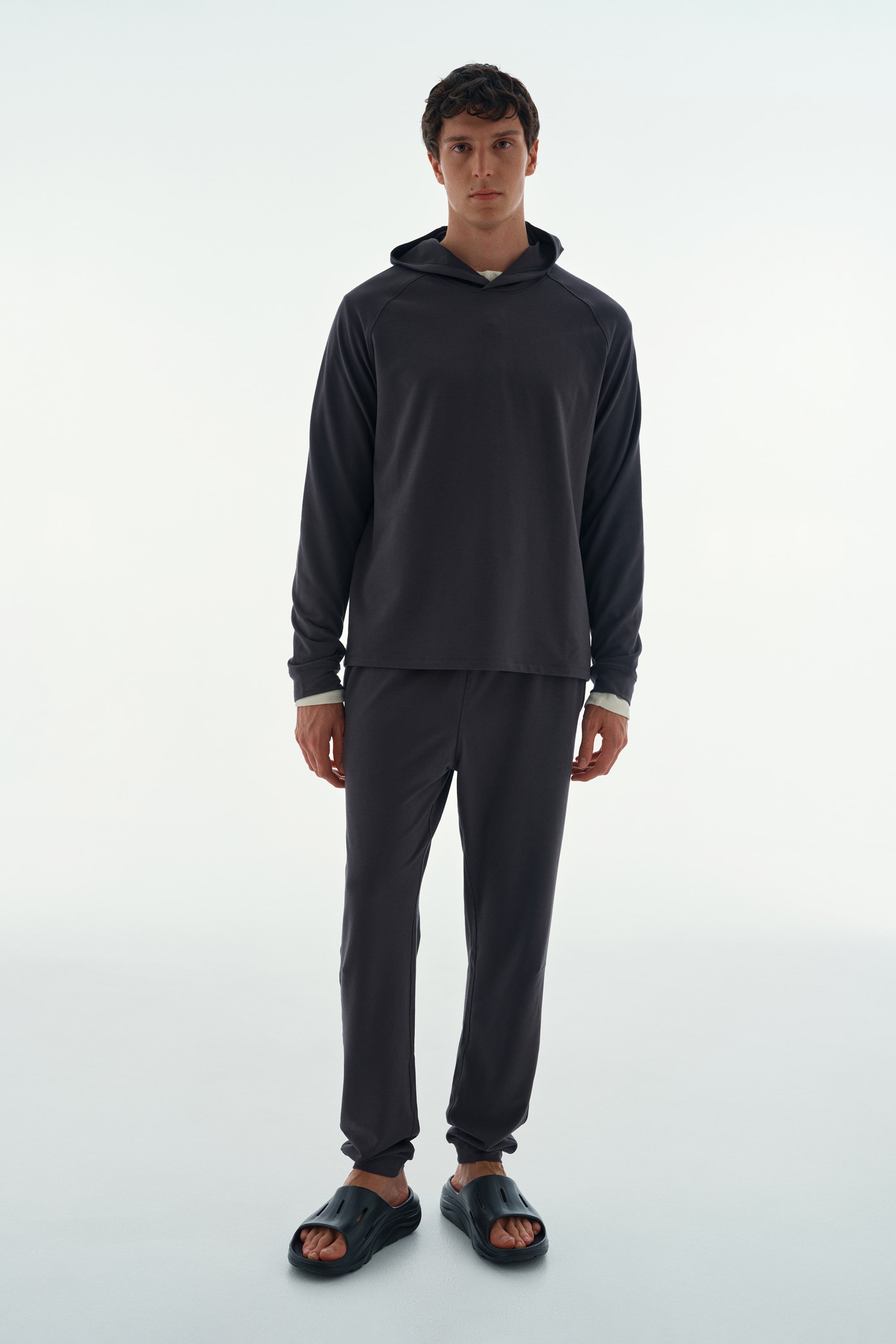 Visualizza l'immagine ingrandita: Un completo loungewear grigio scuro, composto da una felpa con cappuccio a maniche lunghe e pantaloni della tuta coordinati, è indossato dal modello. La felpa presenta maniche raglan sovrapposte a una maglia intima color crema visibile al colletto e ai polsini, mentre i pantaloni dal taglio morbido sono abbinati a sandali slide scuri aperti in punta.